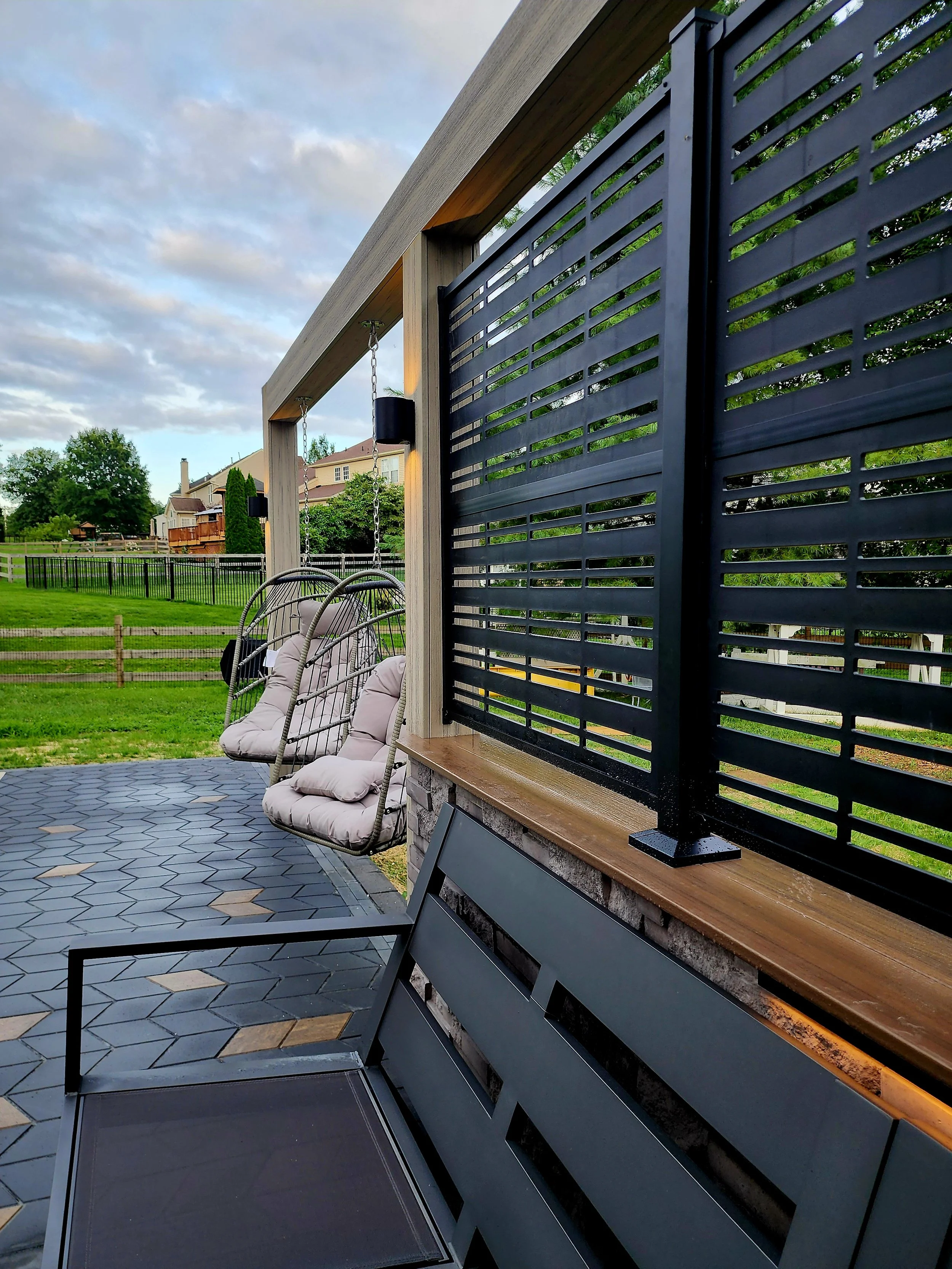 Outdoor patio with two hanging cushioned chairs and a black metal bench, surrounded by a black metal fence and wooden posts, with a grassy yard and houses in the background during cloudy weather.