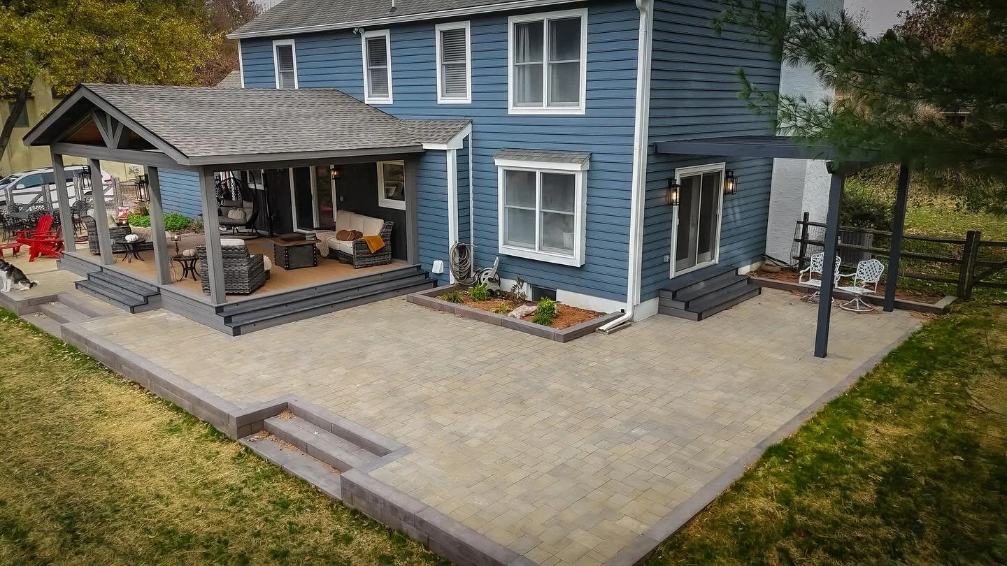 View of a two-story blue house with a large patio area featuring outdoor seating including a covered porch, patio furniture, and potted plants, with a fenced backyard and trees in the background.