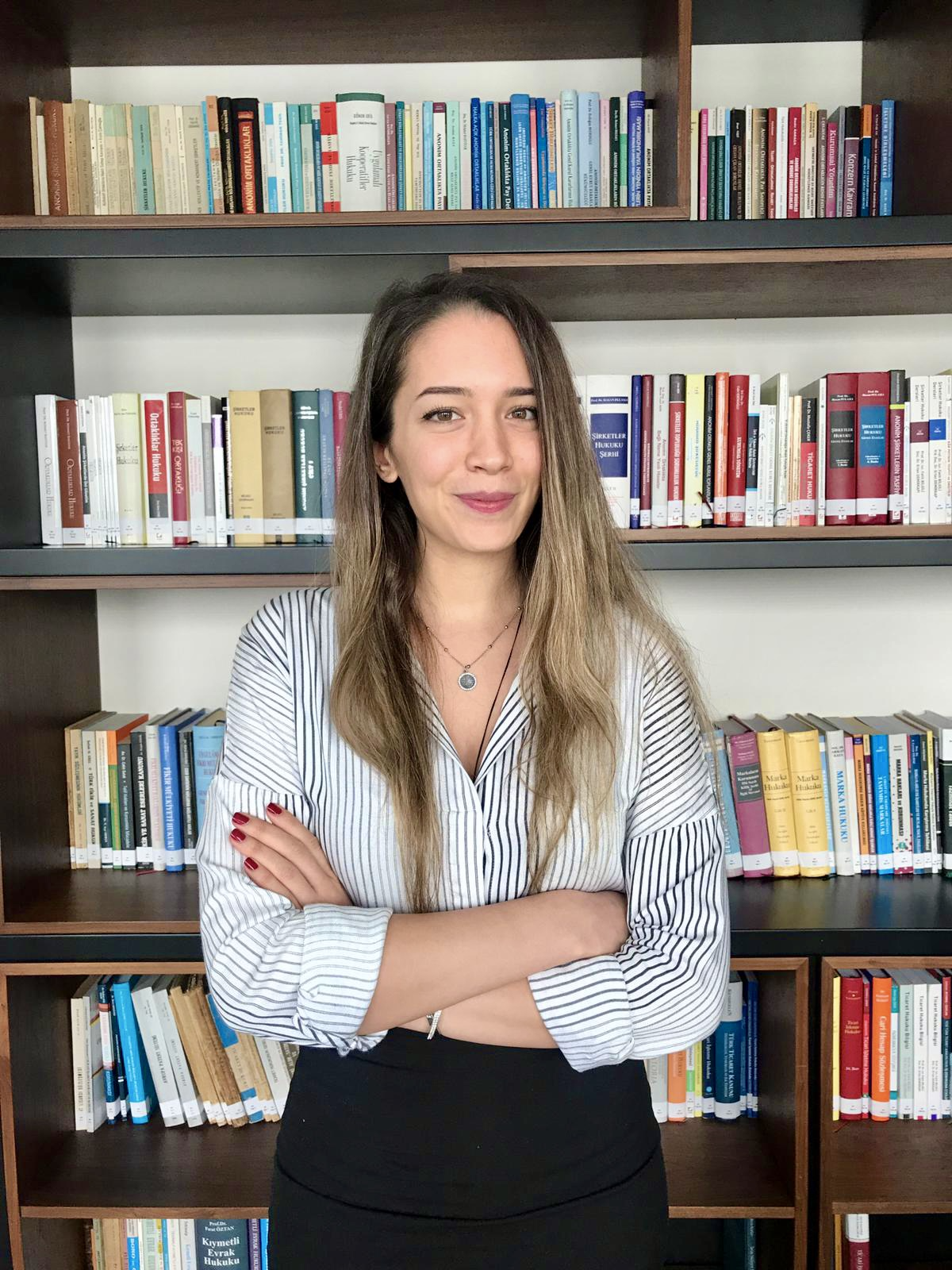 A woman standing in front of a bookshelf filled with books, crossing her arms and smiling at the camera.