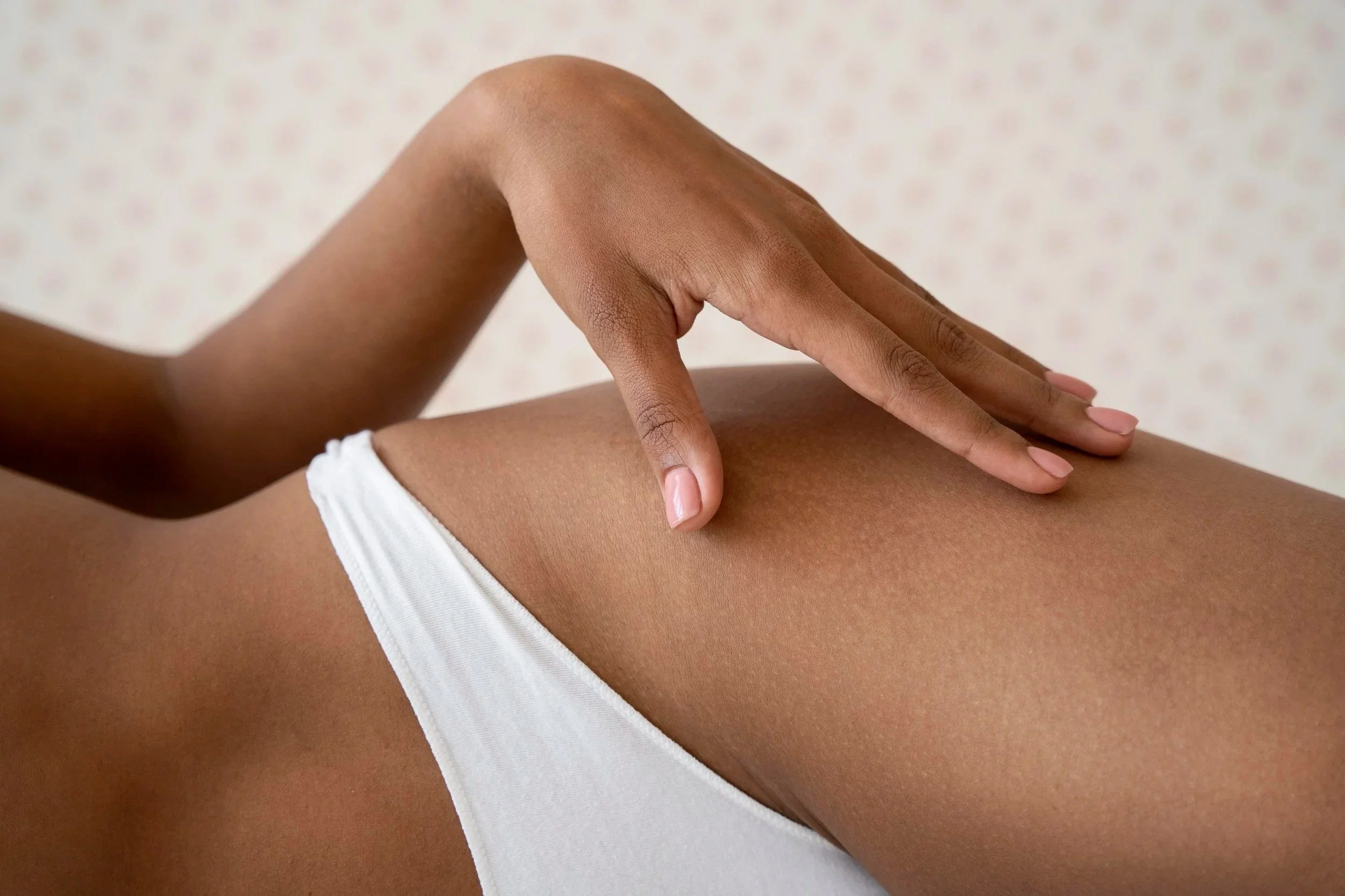 Une main touche la cuisse nue d'une femme portant un sous-vêtement blanc, fond neutre