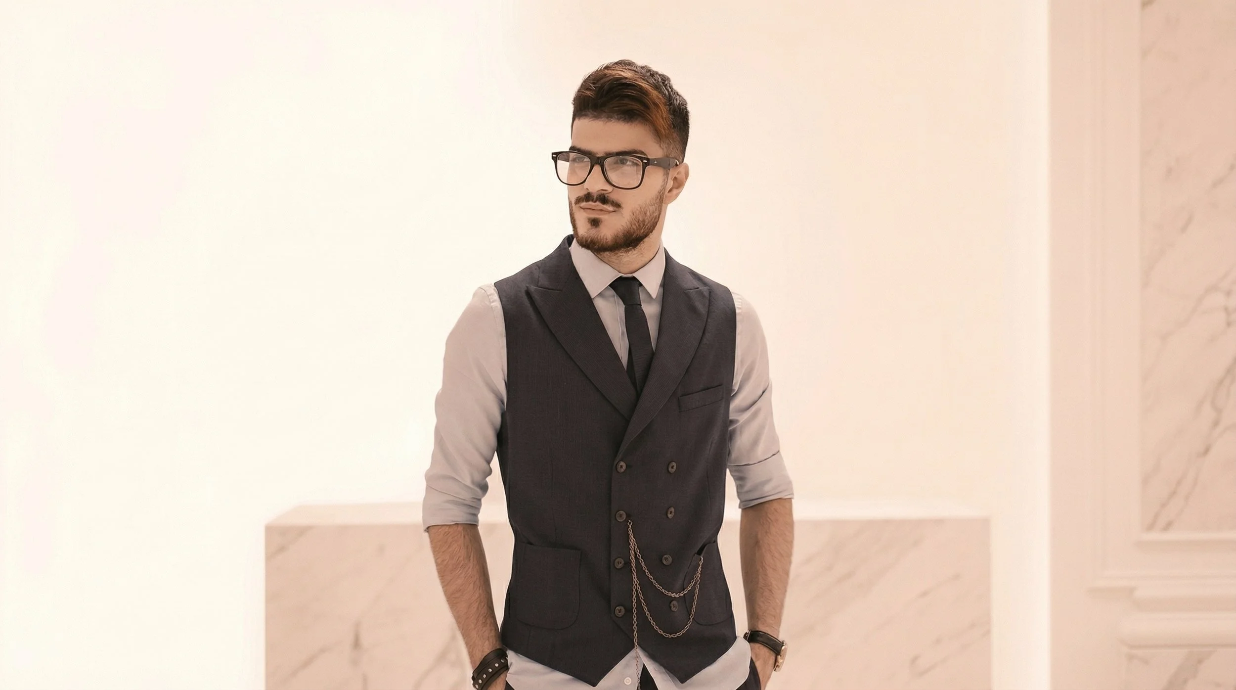Jeune homme portant une veste noire avec chaîne, chemise gris claire, cravate noire, lunettes noires, dans un intérieur avec murs en marbre beige.