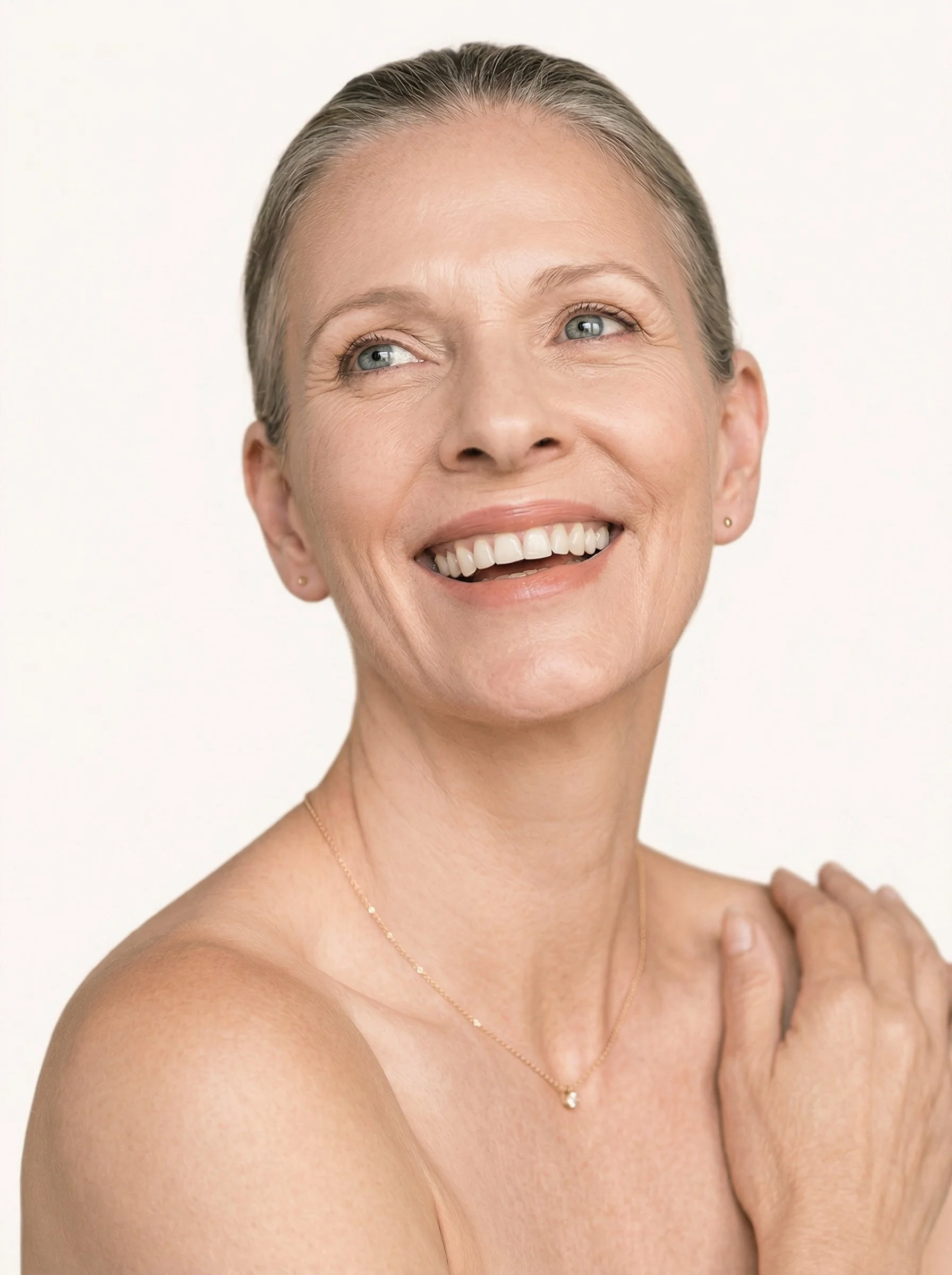 Un portrait d'une femme âgée souriant, avec des cheveux gris, portant une chaîne en or et une petite boucle d'oreille, avec la main sur l'épaule.