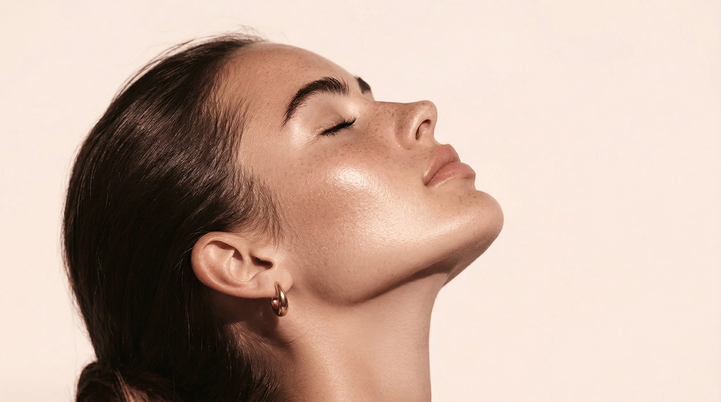 Femme avec peau éclatante, yeux fermés, portant une boucle d'oreille en or, visage de profil, fond clair.
