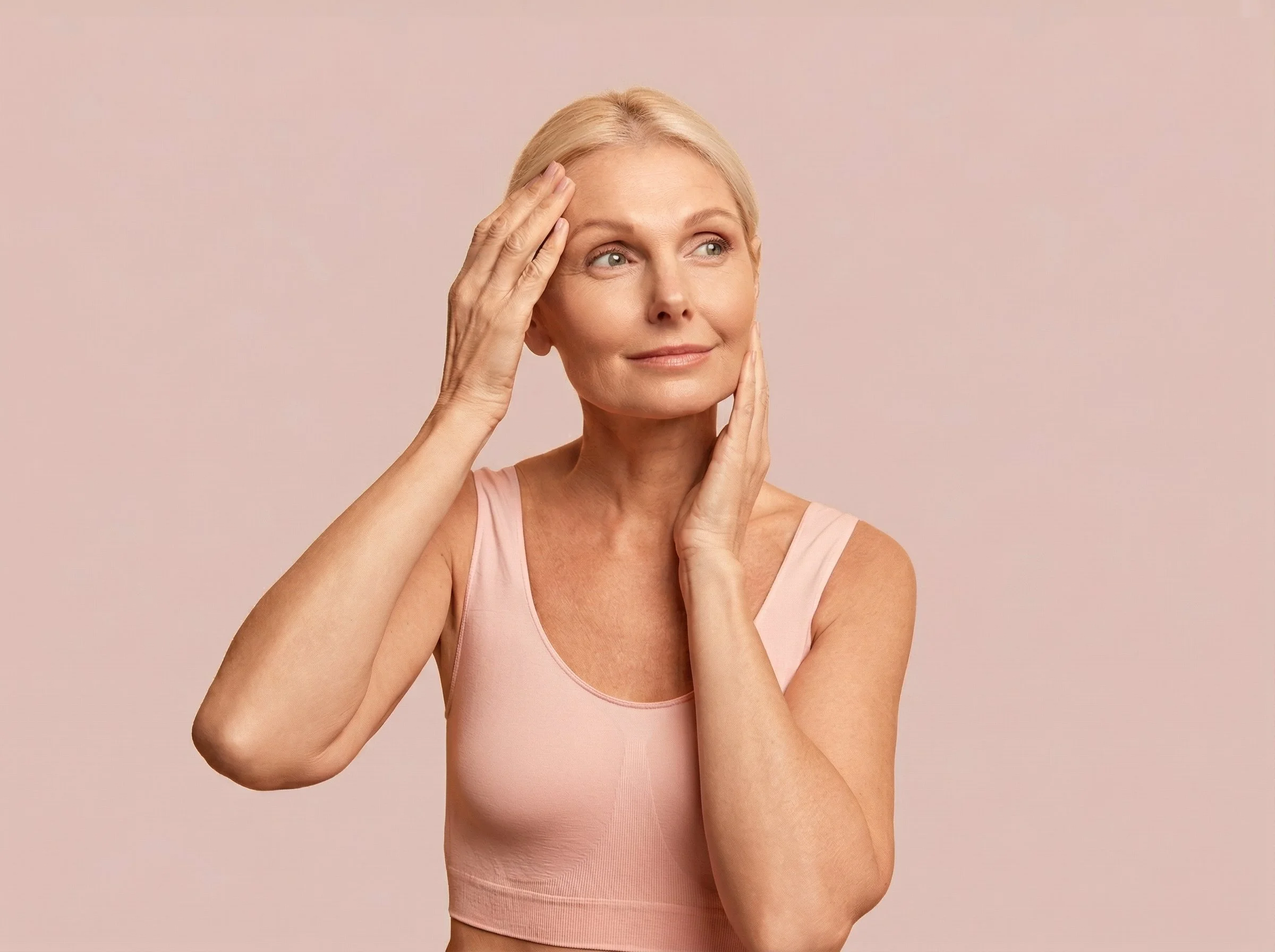 Une femme âgée avec des cheveux blonds courts, portant un haut sans manches rose clair, touche son visage et sa tempe, avec un fond beige clair.