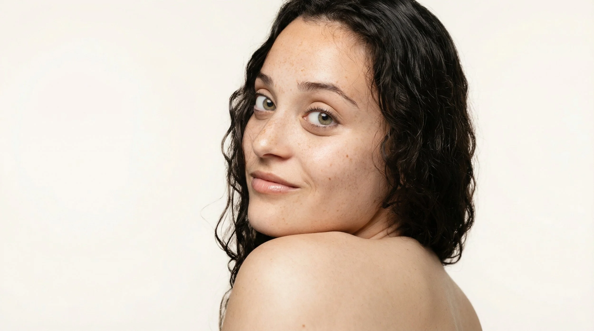Jeune femme avec cheveux bouclés noirs, regardant vers l'objectif, peau claire avec des taches de rousseur, souriant légèrement, sur fond blanc