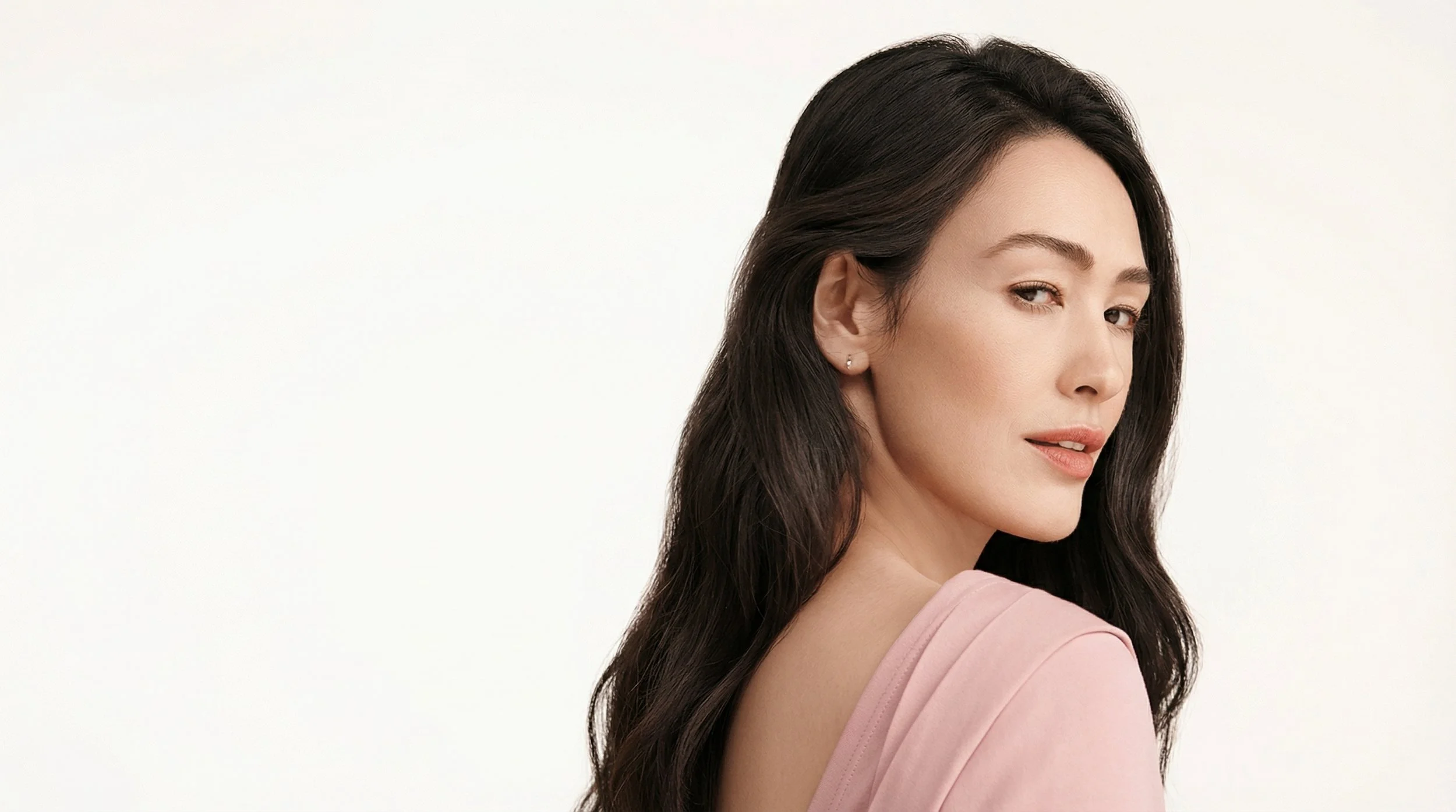 Une femme aux cheveux longs et foncés, portant un vêtement rose pâle, regarde par-dessus son épaule avec un léger sourire, contre un fond blanc.