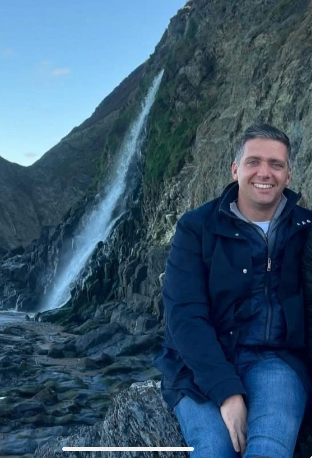 A man sitting on rocks near a mountain with a waterfall in the background, smiling at the camera.