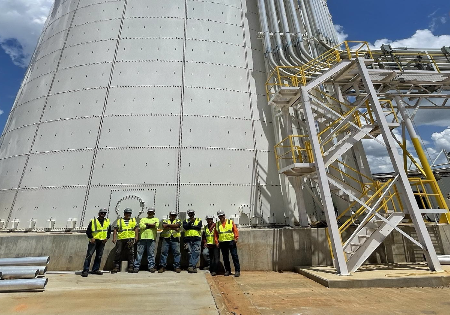 A group of workers in safety vests and helmets standing in front of a large industrial tank or silo with scaffolding and pipes.