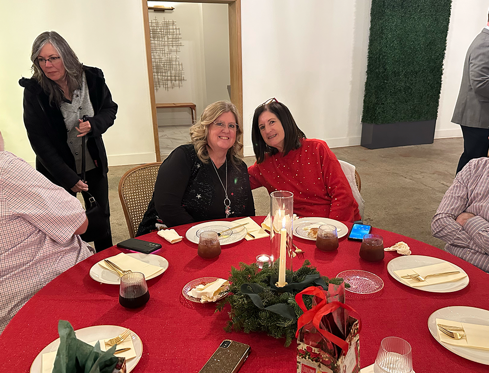 Two women smiling and sitting at a holiday dinner table decorated with a candle centerpiece and greenery, with other guests in the background.