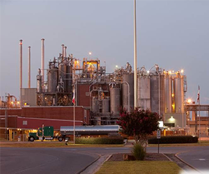Refinery with large industrial structures and pipes, illuminated at dusk or night.