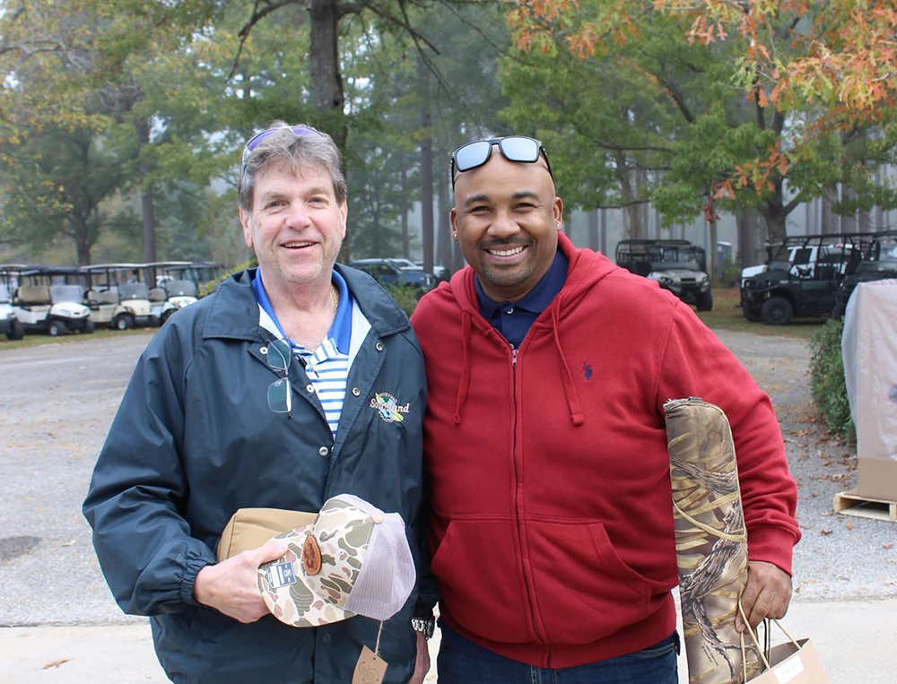 Two smiling men standing outdoors in a parking lot surrounded by trees with autumn leaves. One man is wearing a navy jacket, glasses, and holding a cap, while the other man is wearing a red hoodie and sunglasses on his head, holding a camouflage-patterned item.