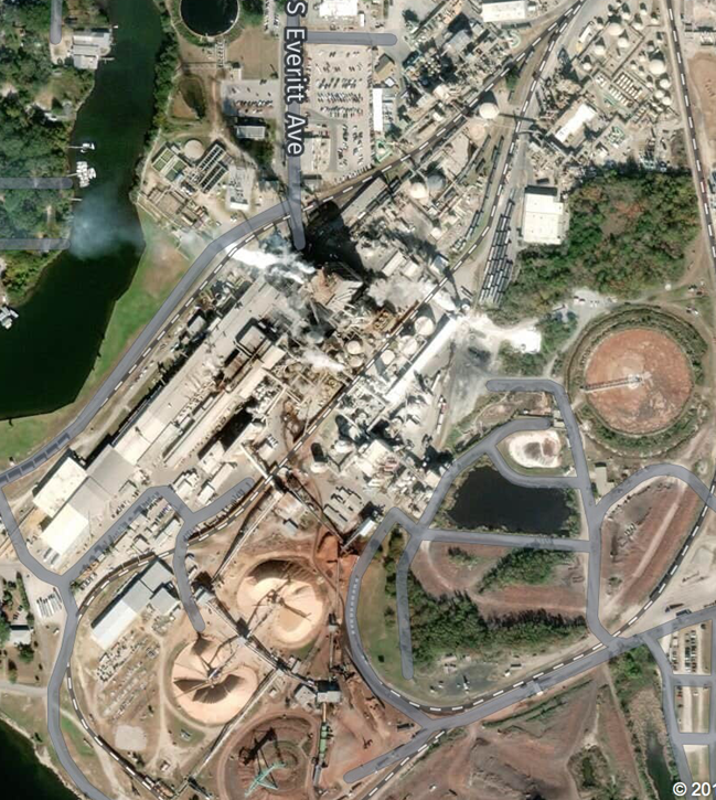 Aerial view of an industrial site with multiple buildings, access roads, piping, and storage tanks, located near a body of water and a road labeled 'S Eventt Ave.'