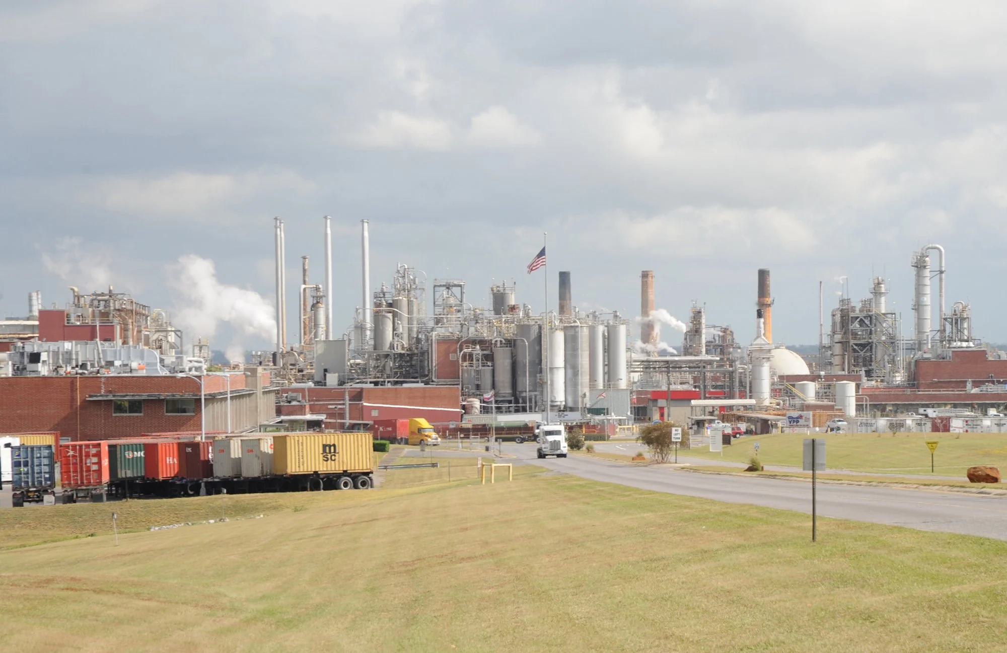 Industrial factory with multiple smokestacks emitting smoke or steam, American flag flying, train with containers on tracks outside the facility, and well-maintained grassy area in the foreground.