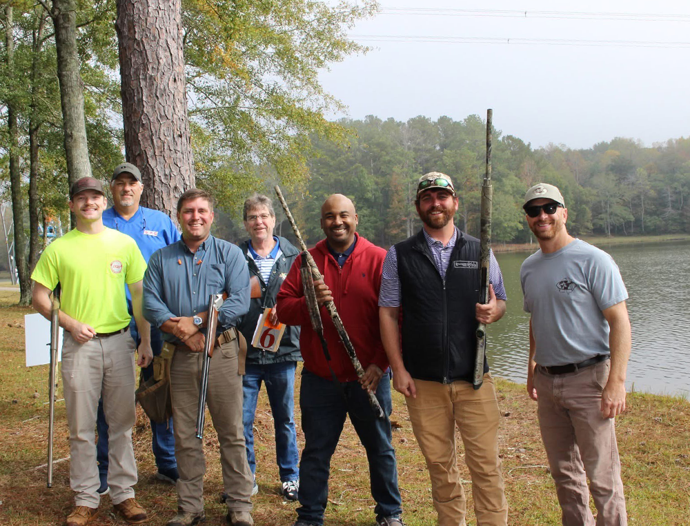 A group of seven men standing outdoors near a lake, some holding rifles, with trees and a partly cloudy sky in the background.