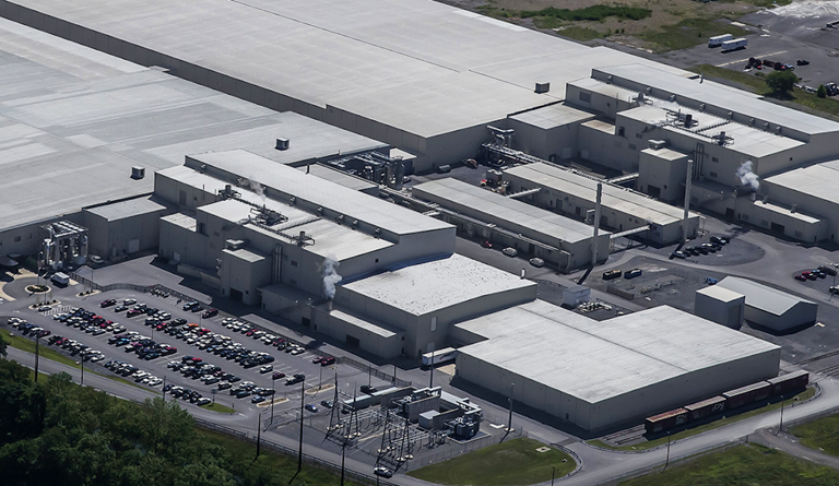 Aerial view of a large industrial or manufacturing facility with multiple connected buildings, parking lots filled with cars, and utility structures.