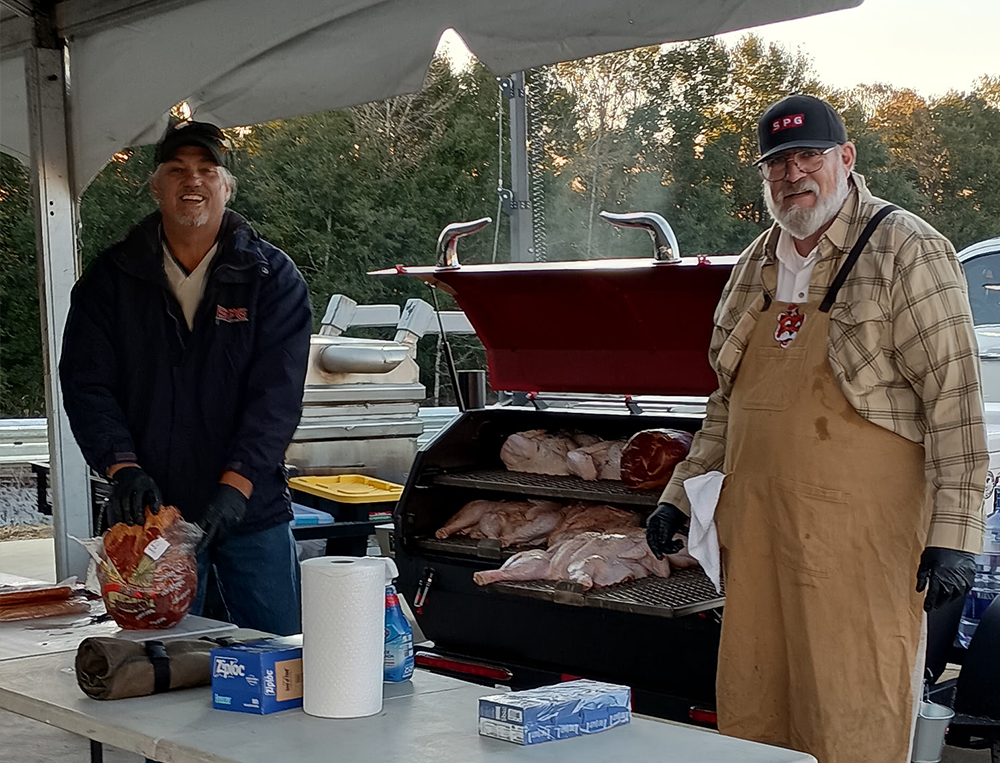 Two men cooking and preparing meat, including ribs and brisket, at an outdoor barbecue event under a tent.