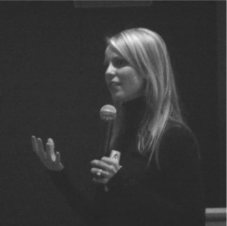 A woman with blonde hair holding a microphone, gesturing with her other hand, in a dark setting.