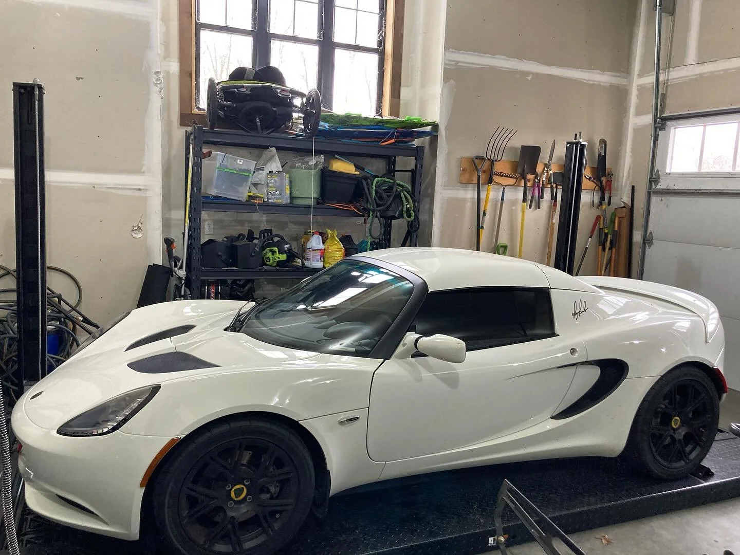 A white Lotus sports car parked inside a garage with tools and equipment on shelves and hanging on the wall.