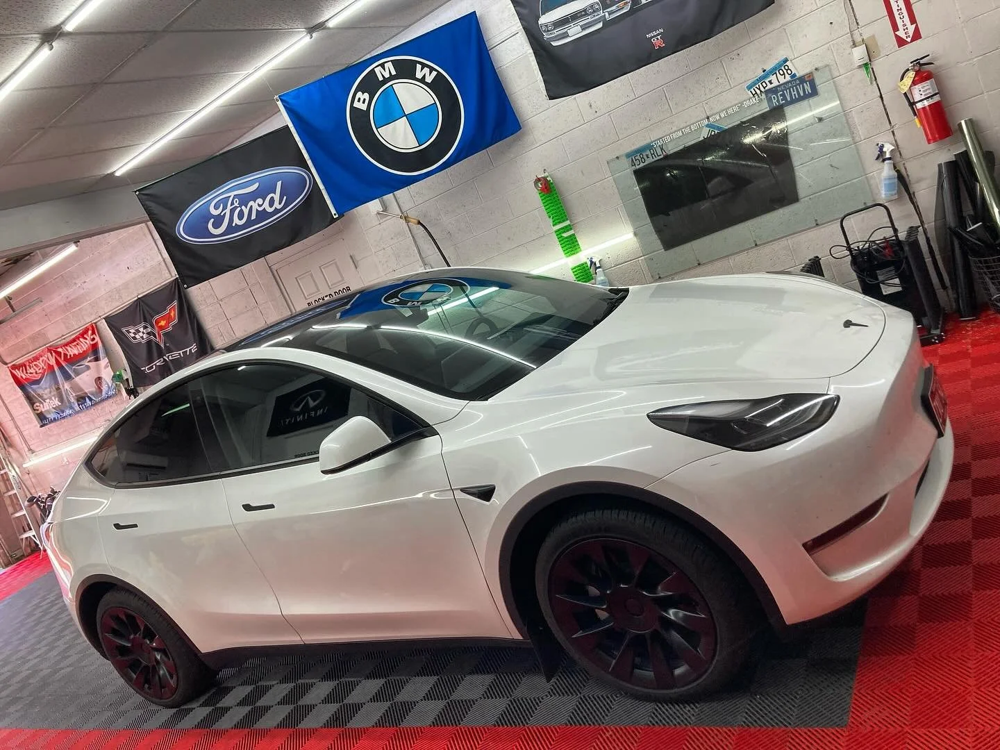 A white Tesla parked in a dealership or showroom with automotive banners and flags of Ford, BMW, Corvette, and Infiniti on the wall.