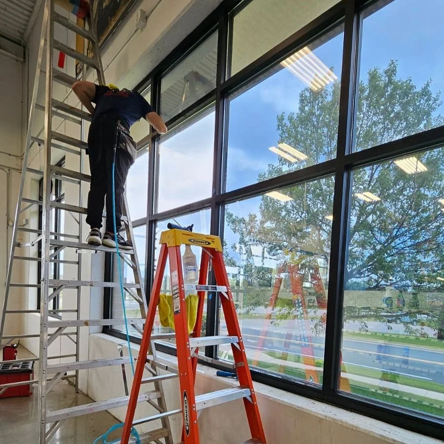 A worker standing on a ladder cleaning large window panes in a commercial building.