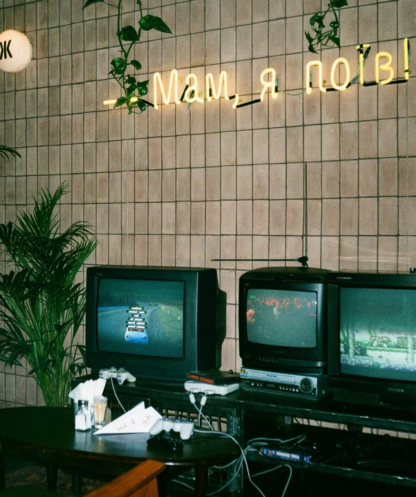 Interior of a room with a beige tiled wall, a neon sign, vintage televisions, a potted plant, and gaming consoles.