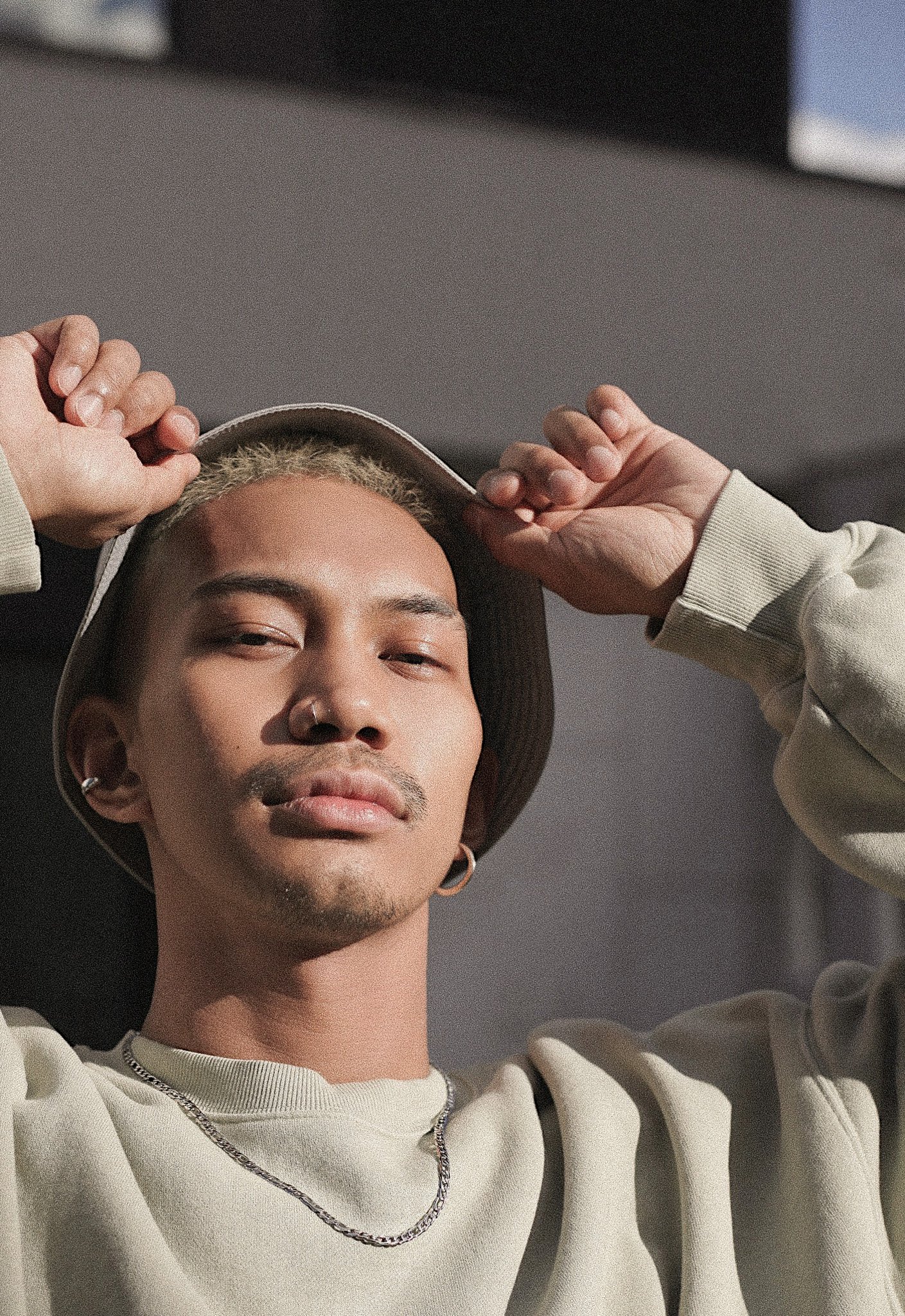 A young person in street fashion style, wearing hoop earrings is wearing a beige sweatshirt and chain necklace, adjusting a bucket hat with both hands, looking confidently into the camera in a close-up shot.