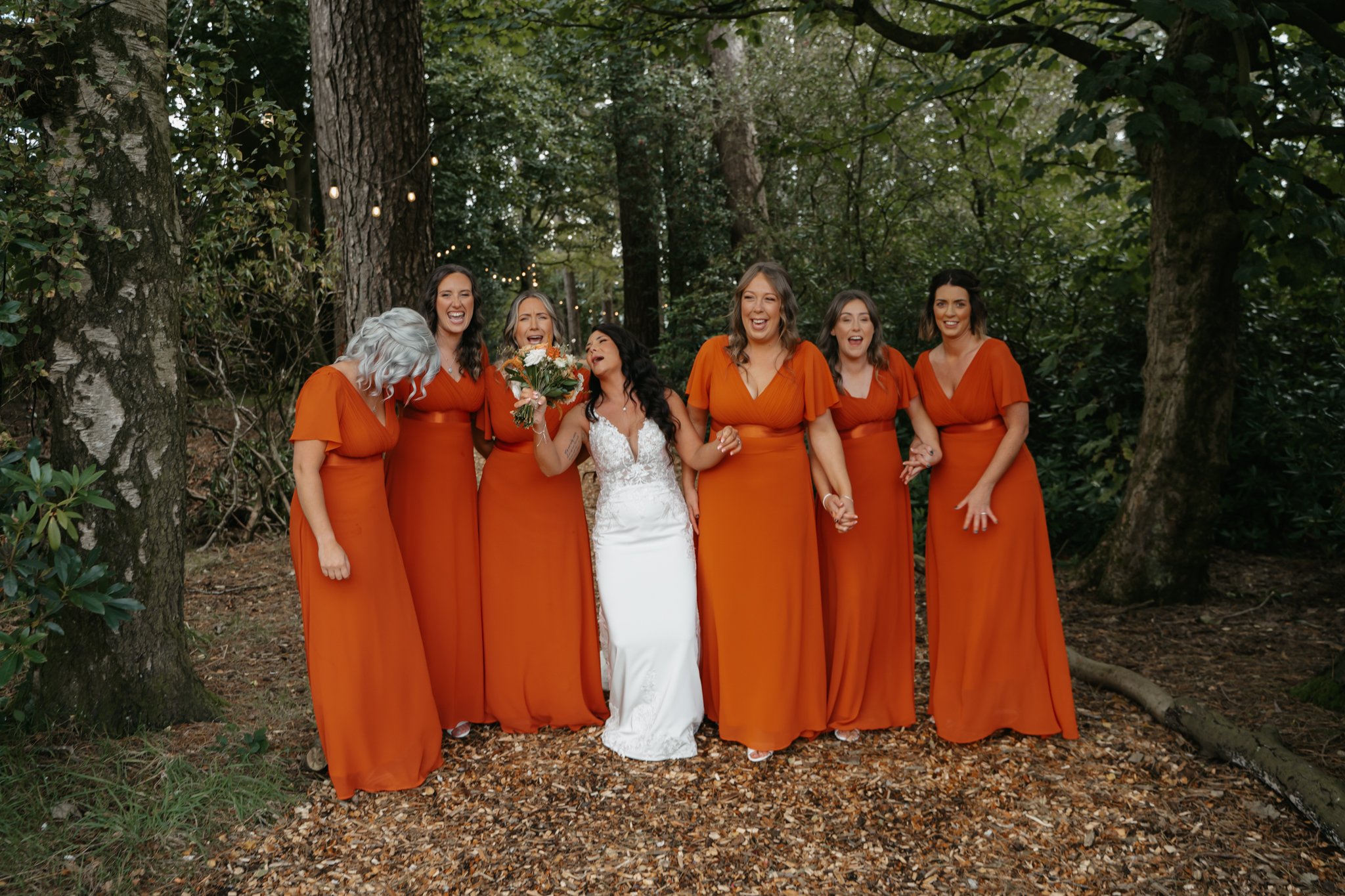 Bride in a white wedding dress holding a bouquet of flowers walking with six bridesmaids dressed in matching burnt orange dresses in a wooded outdoor setting.