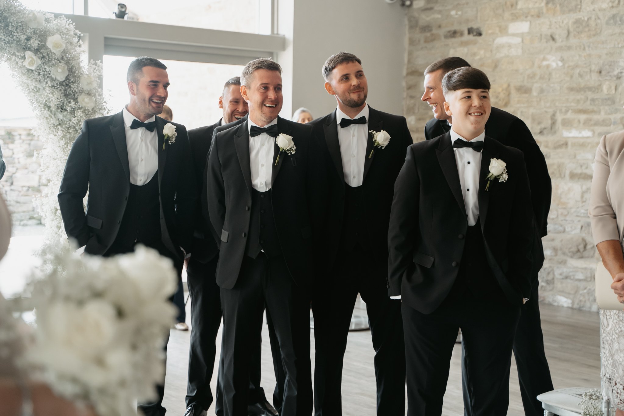 A group of men in black tuxedos with white shirts and black bow ties, smiling and standing together at a wedding ceremony.