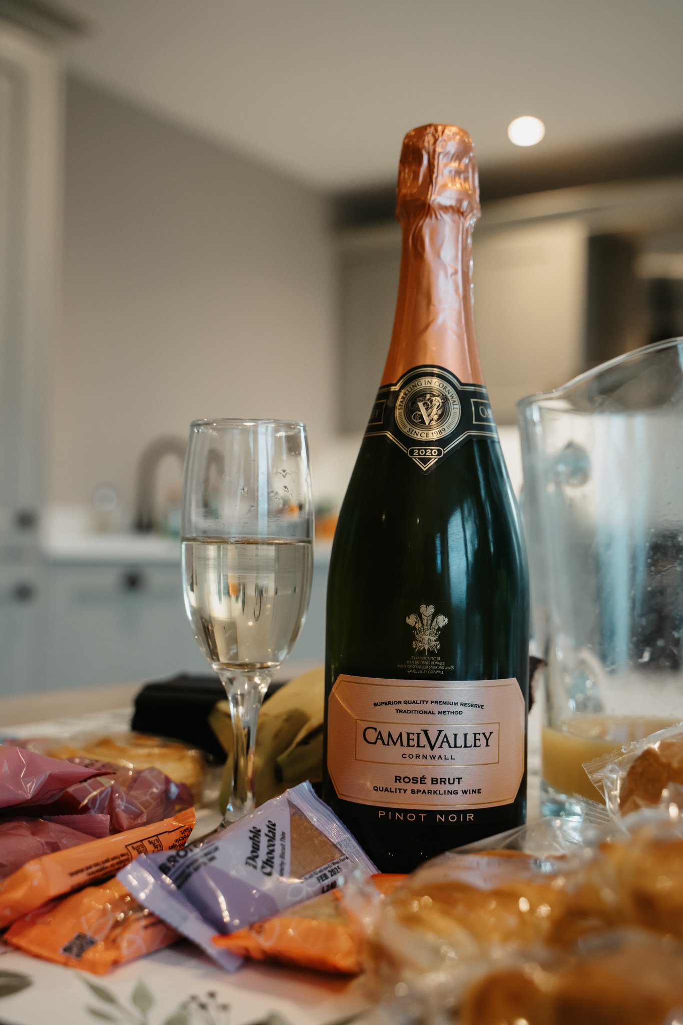 A bottle of Camel Valley Rosé Brut sparkling wine with a glass of champagne on a cluttered table, surrounded by snacks and food in a home kitchen setting.