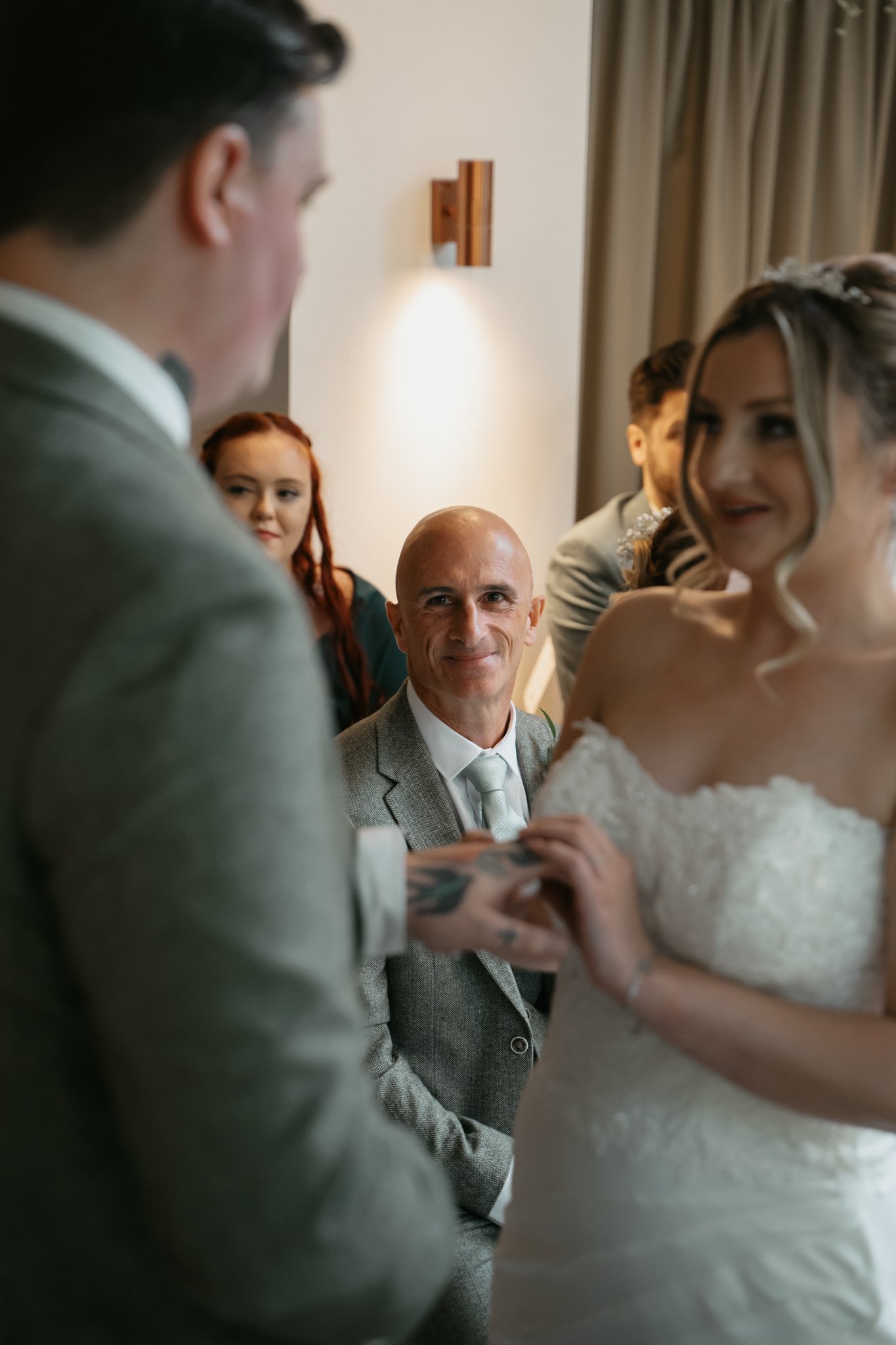 Bald man in a gray suit smiling during a wedding ceremony, holding hands with a bride in a strapless lace wedding dress, with a woman with red hair and a man with dark hair in the background.