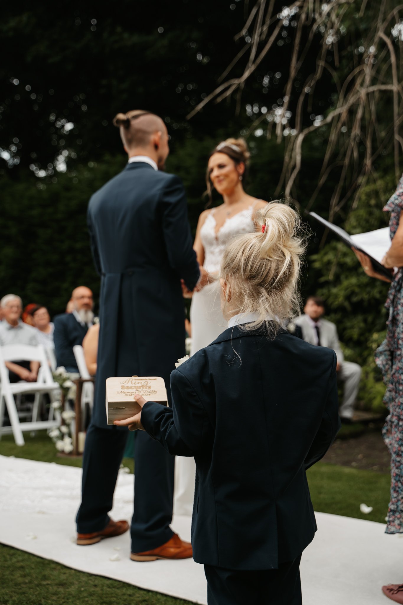 A young girl holding a wooden box with wedding vows during an outdoor wedding ceremony, with a bride and groom exchanging vows in the background.