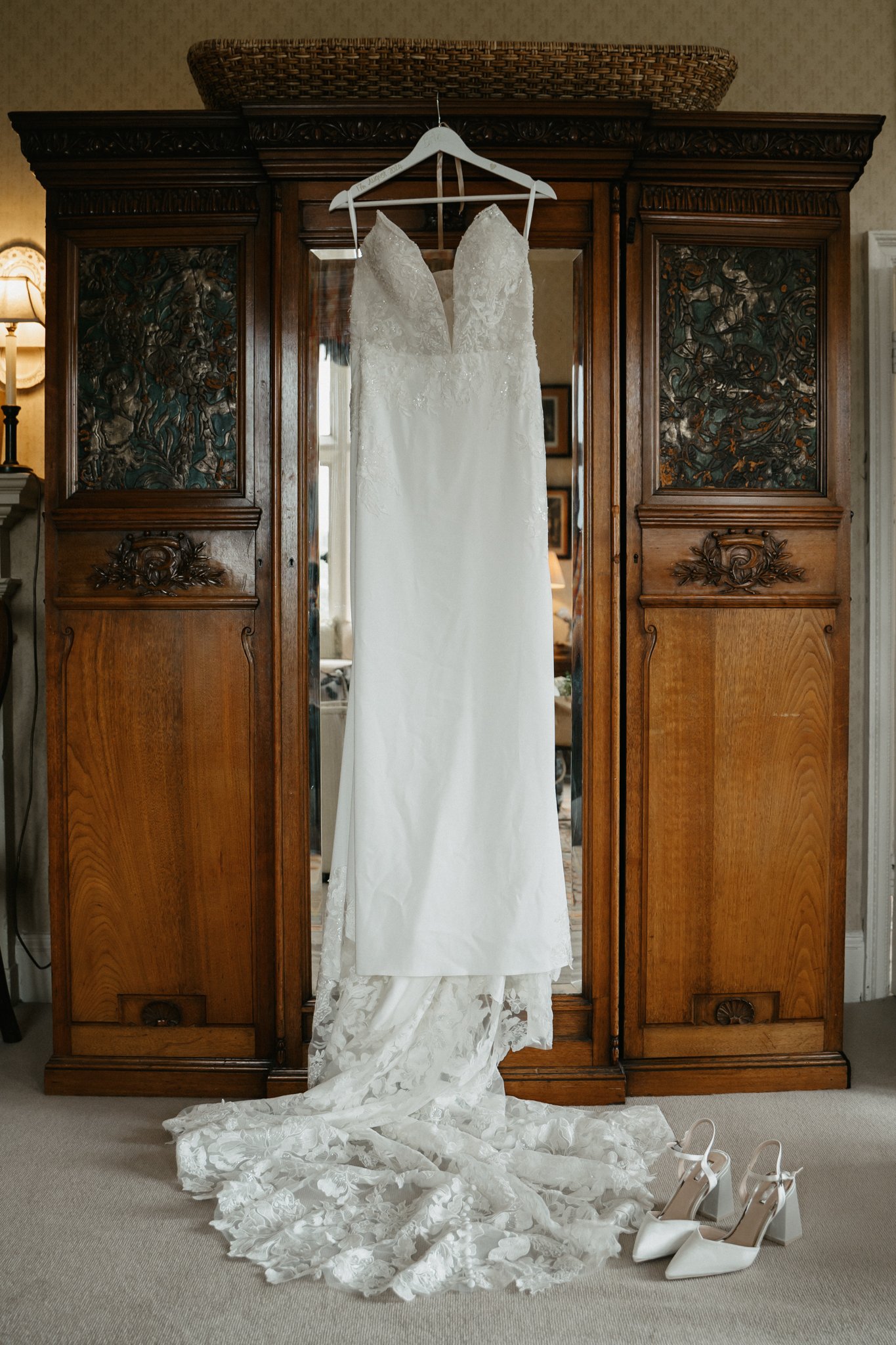 White wedding dress hanging on a wooden wardrobe with a pair of white high heels on the floor below.