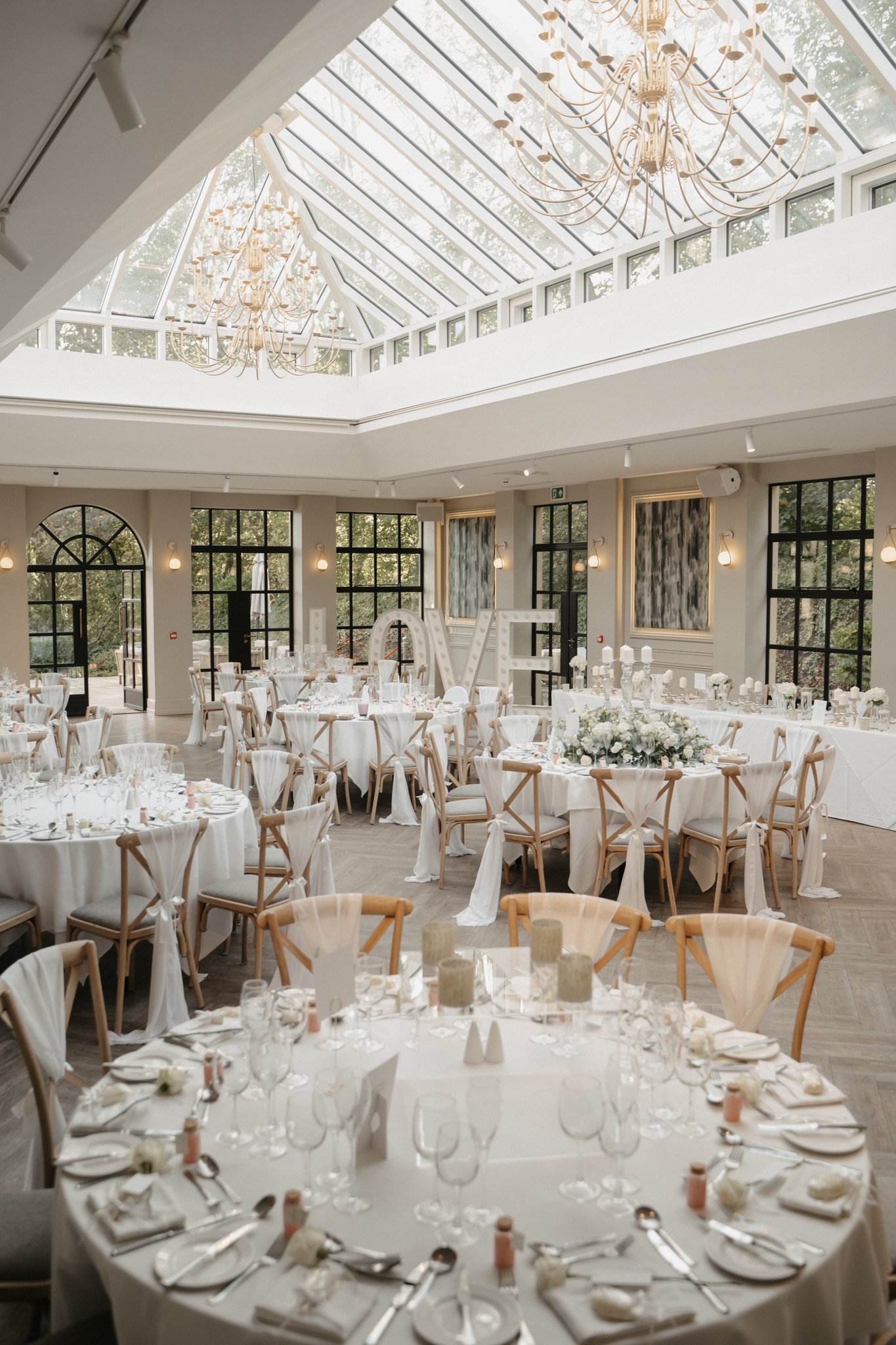 Elegant wedding reception hall with round tables covered in white tablecloths, decorated with white floral centerpieces and candles, wooden chairs with white fabric bows, large windows, a glass ceiling with chandeliers, and a large illuminated love s