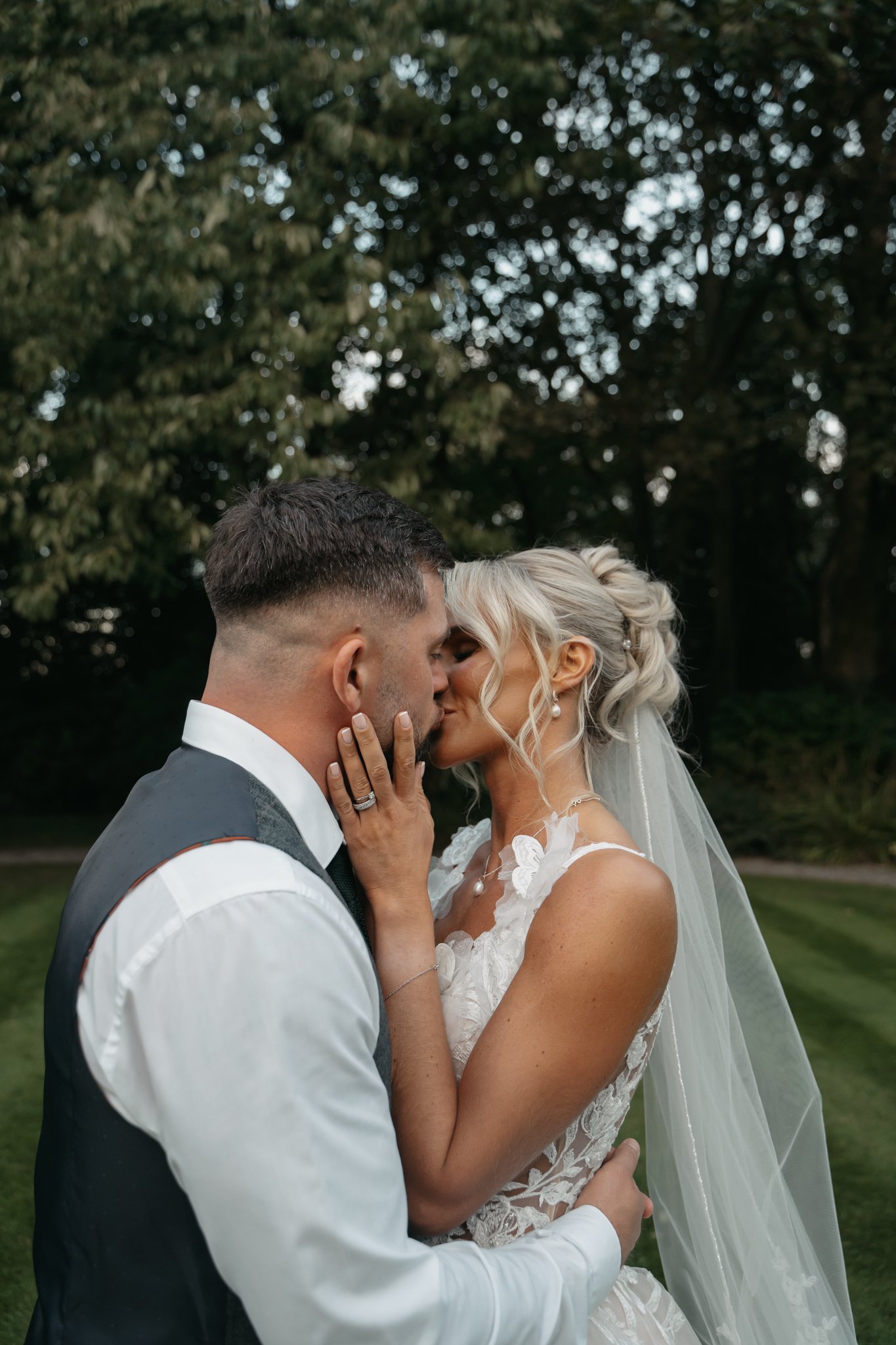 A bride and groom kissing outdoors in a garden, with the groom wearing a vest and tie and the bride wearing a white lace wedding dress and veil.