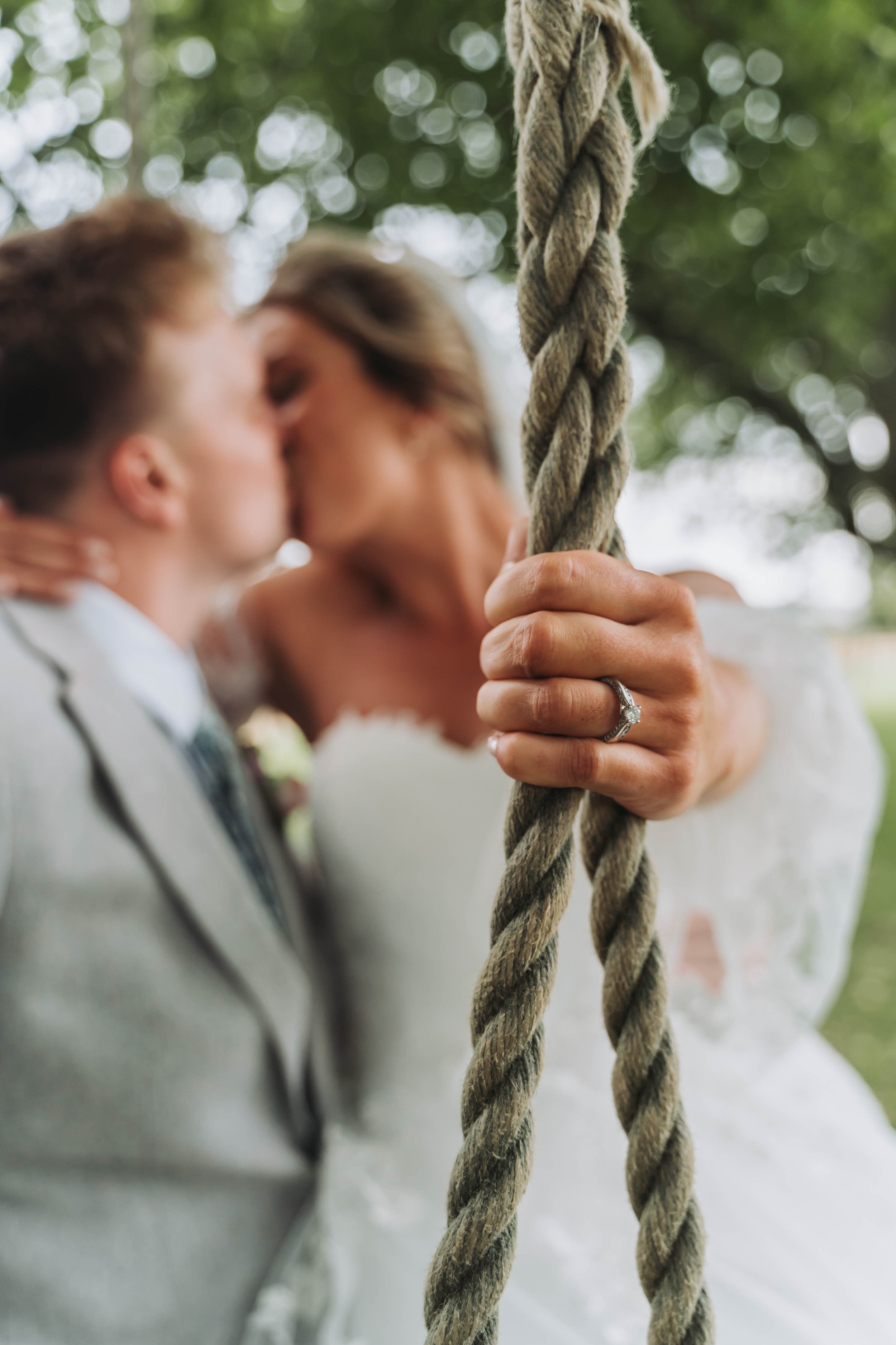 A bride and groom kissing behind a thick rope swing, with the bride's hand featuring a wedding ring holding the rope.