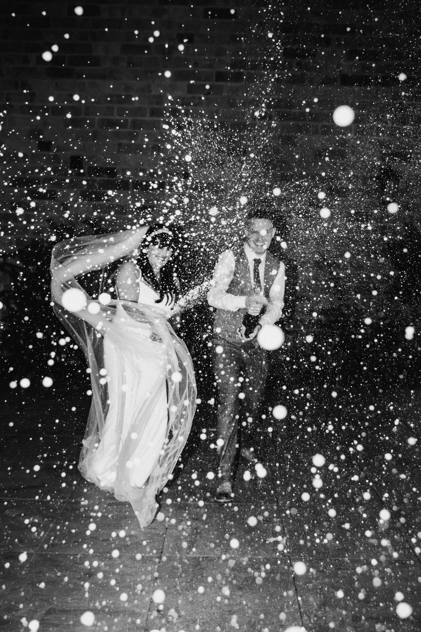 A couple at a wedding or celebration dancing and being splashed with confetti or spray. The scene is lively with motion and joyful expressions.