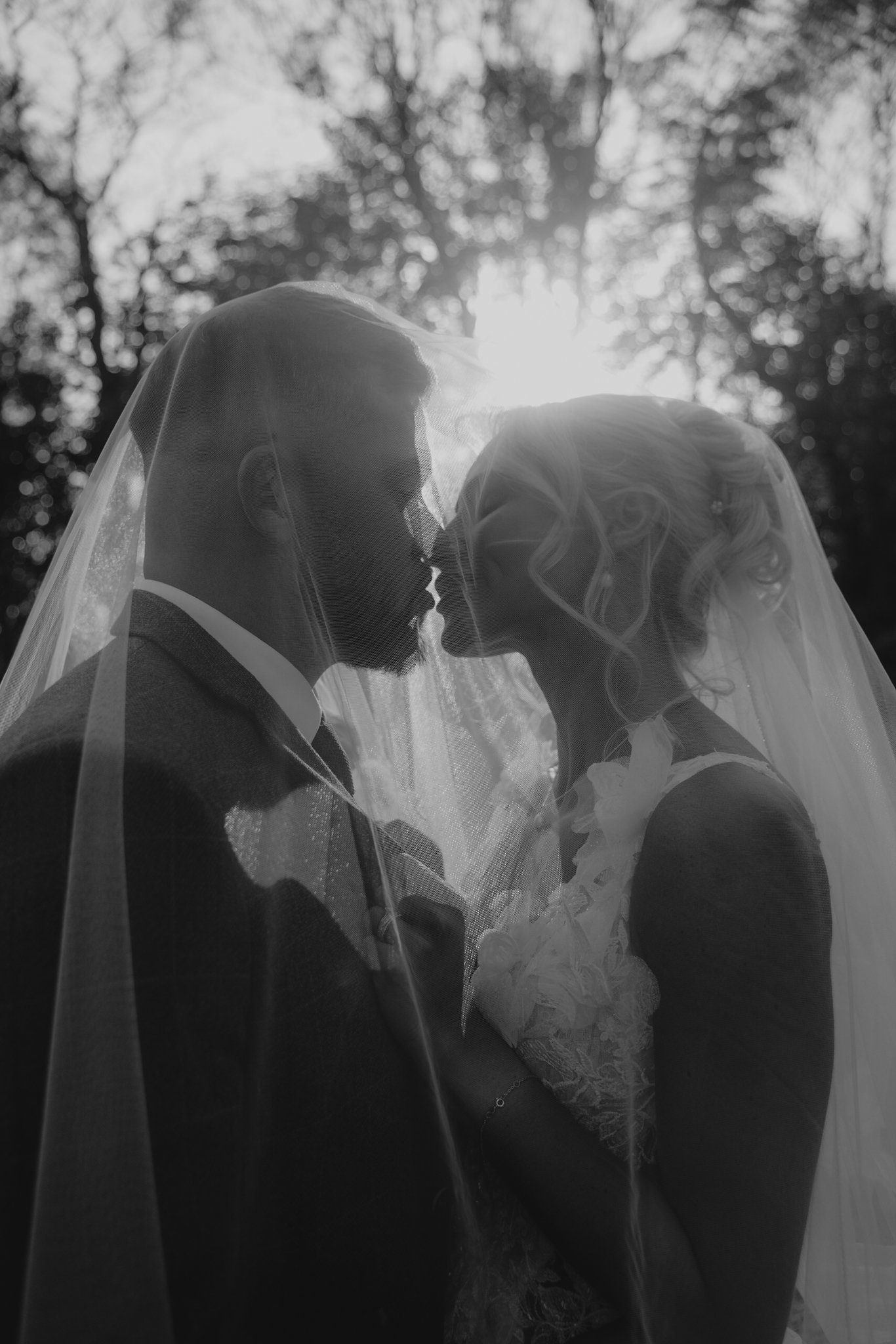 Black and white photograph of a bride and groom facing each other with their noses touching, their faces partially obscured by a veil, outdoors with sunlight and trees in the background.