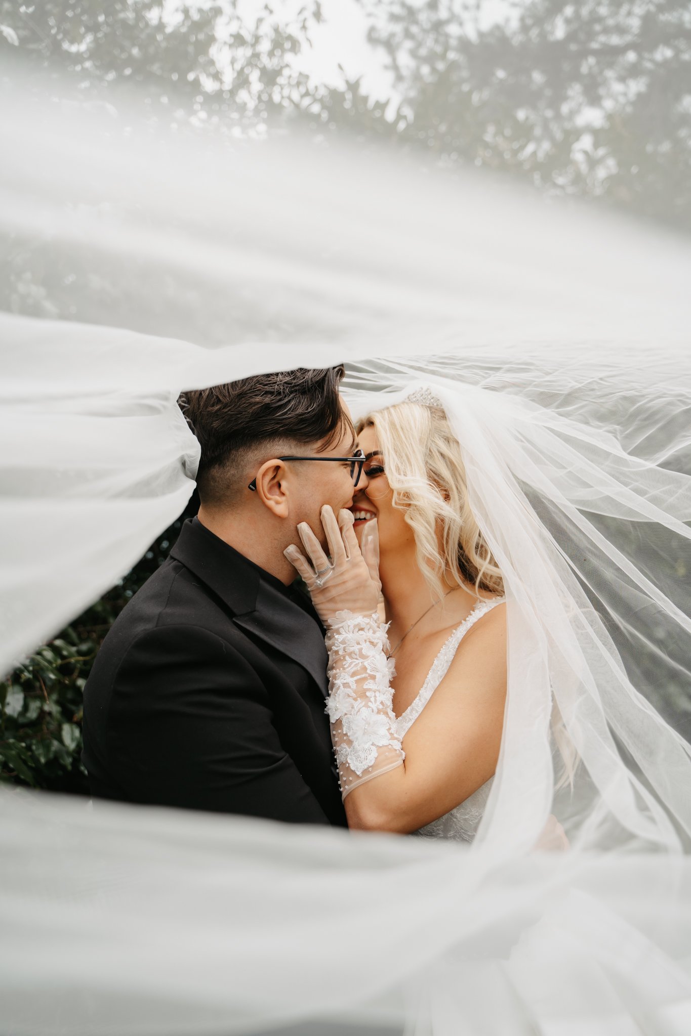 A bride and groom sharing a kiss and smiling, framed by the bride's veil and outdoors with trees in the background.