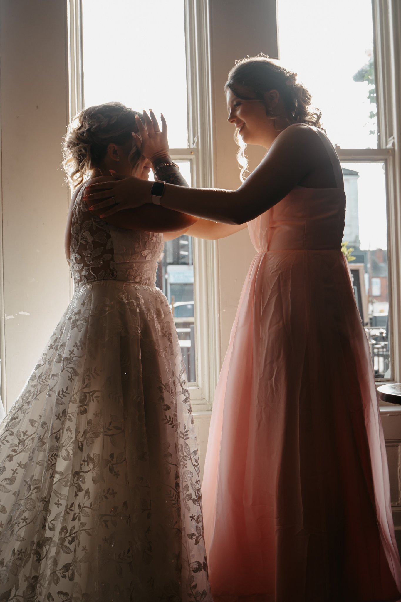 Two women, one in a wedding dress, emotionally embrace in front of a bright window.