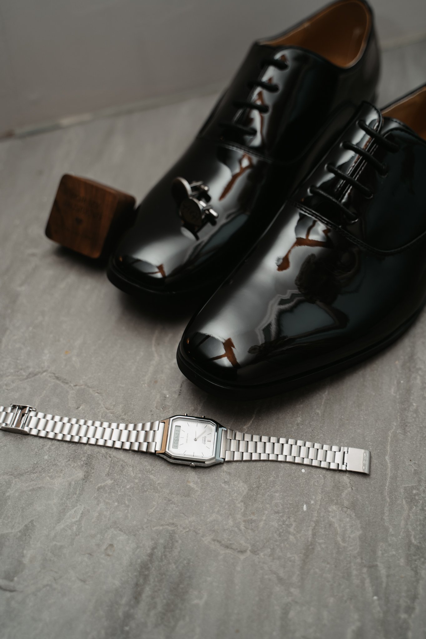 A pair of shiny black leather dress shoes, a silver wristwatch with a white face, and a skateboard wheel on a gray surface.