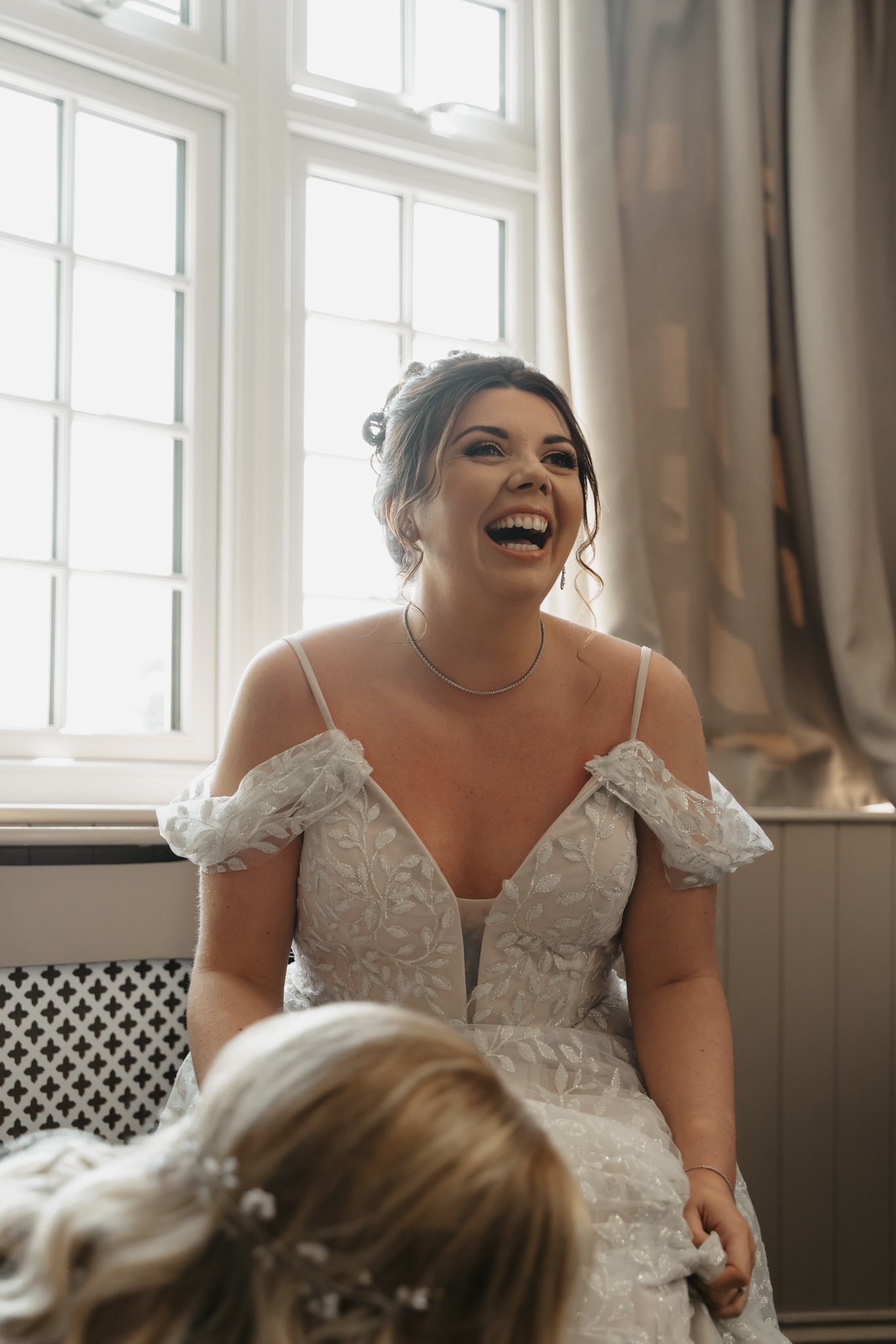 A woman dressed in a wedding gown laughing joyfully inside a room with large windows and curtains.