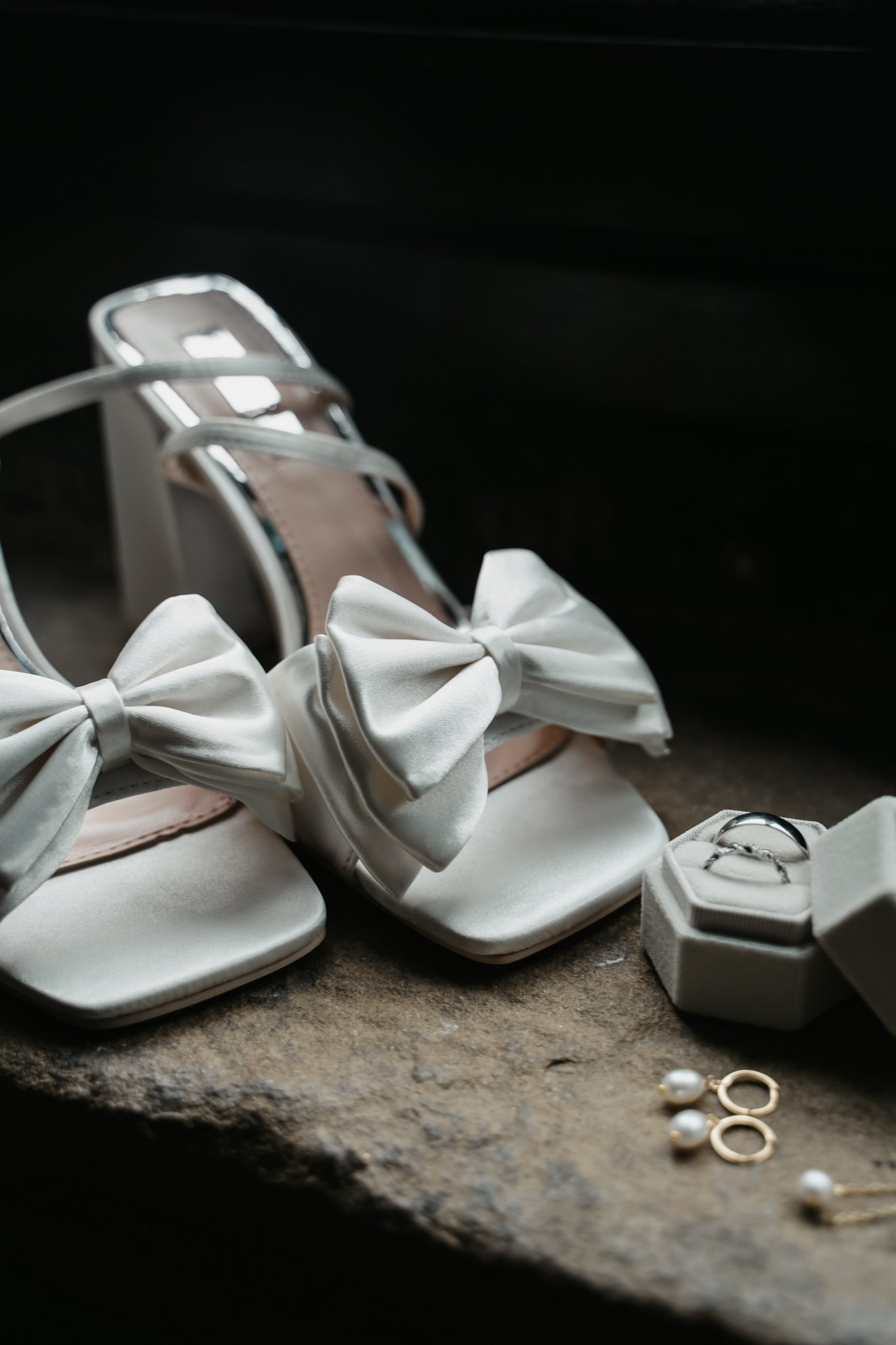 A pair of white satin wedding shoes with bows, a small box with wedding rings, and pearl and gold earrings on a rustic surface.