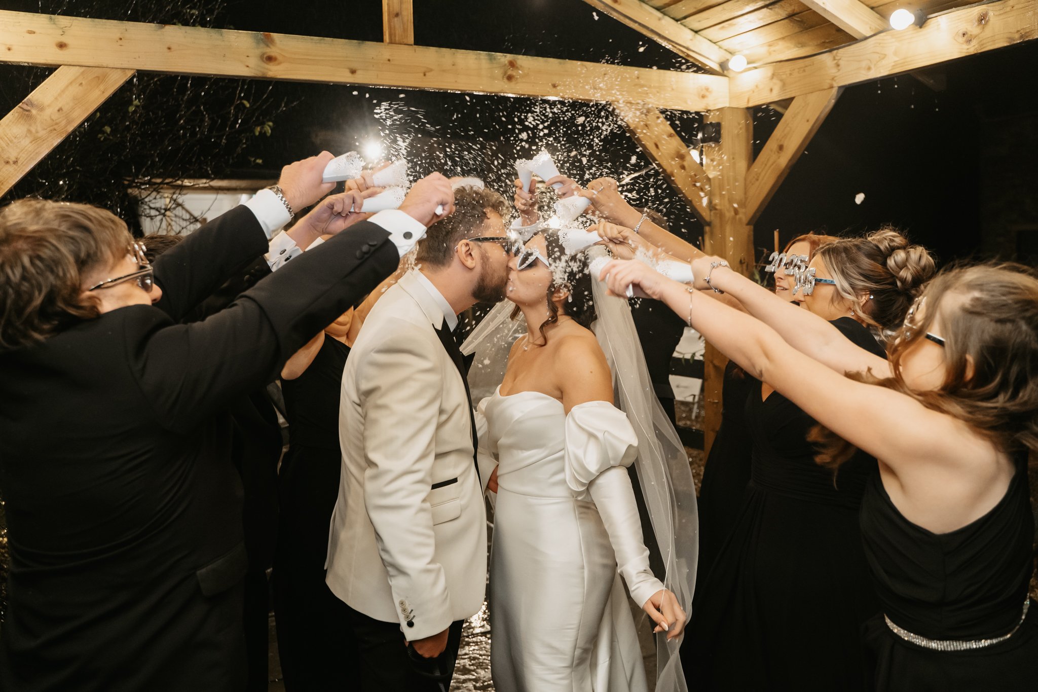 Bride and groom kissing under a wooden wedding arch while being showered with confetti by friends and family at night.