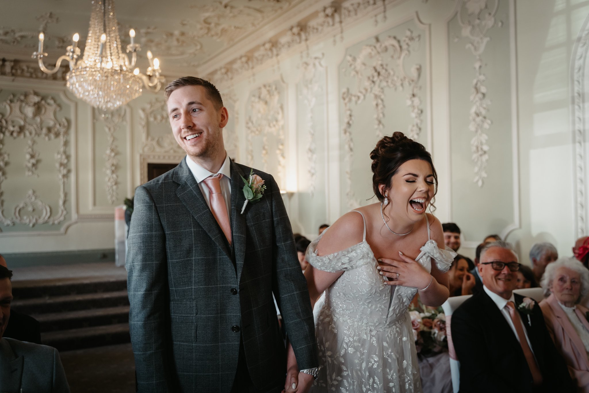 A bride and groom holding hands and laughing during their wedding ceremony in an elegant, ornately decorated room with a chandelier.