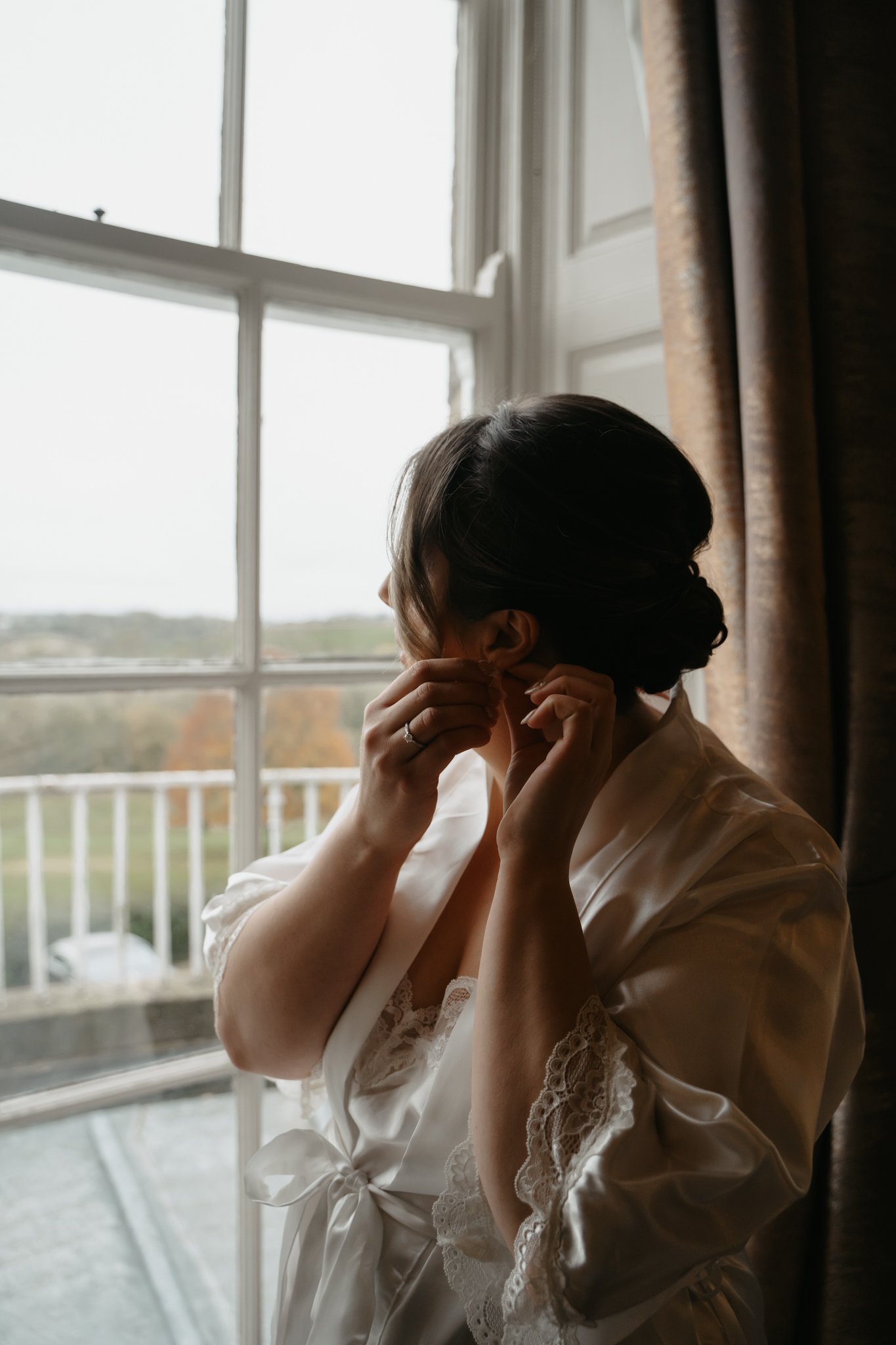 A woman in a satin robe with lace trim adjusting her earrings near a window