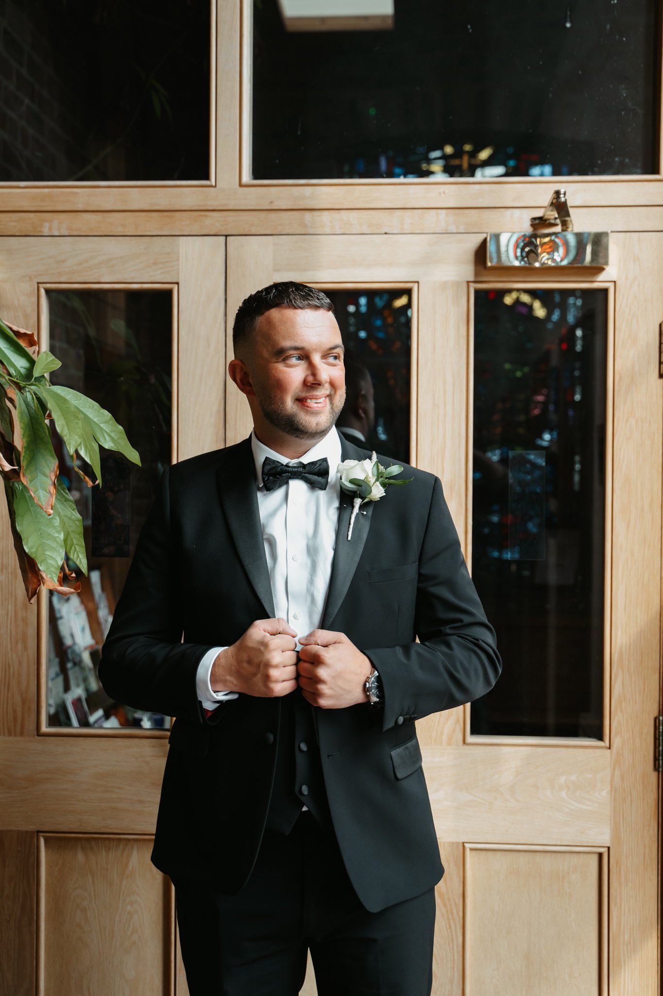 A groom in a black tuxedo with a white shirt and black bow tie, standing in front of a wooden door, smiling and looking to the side.