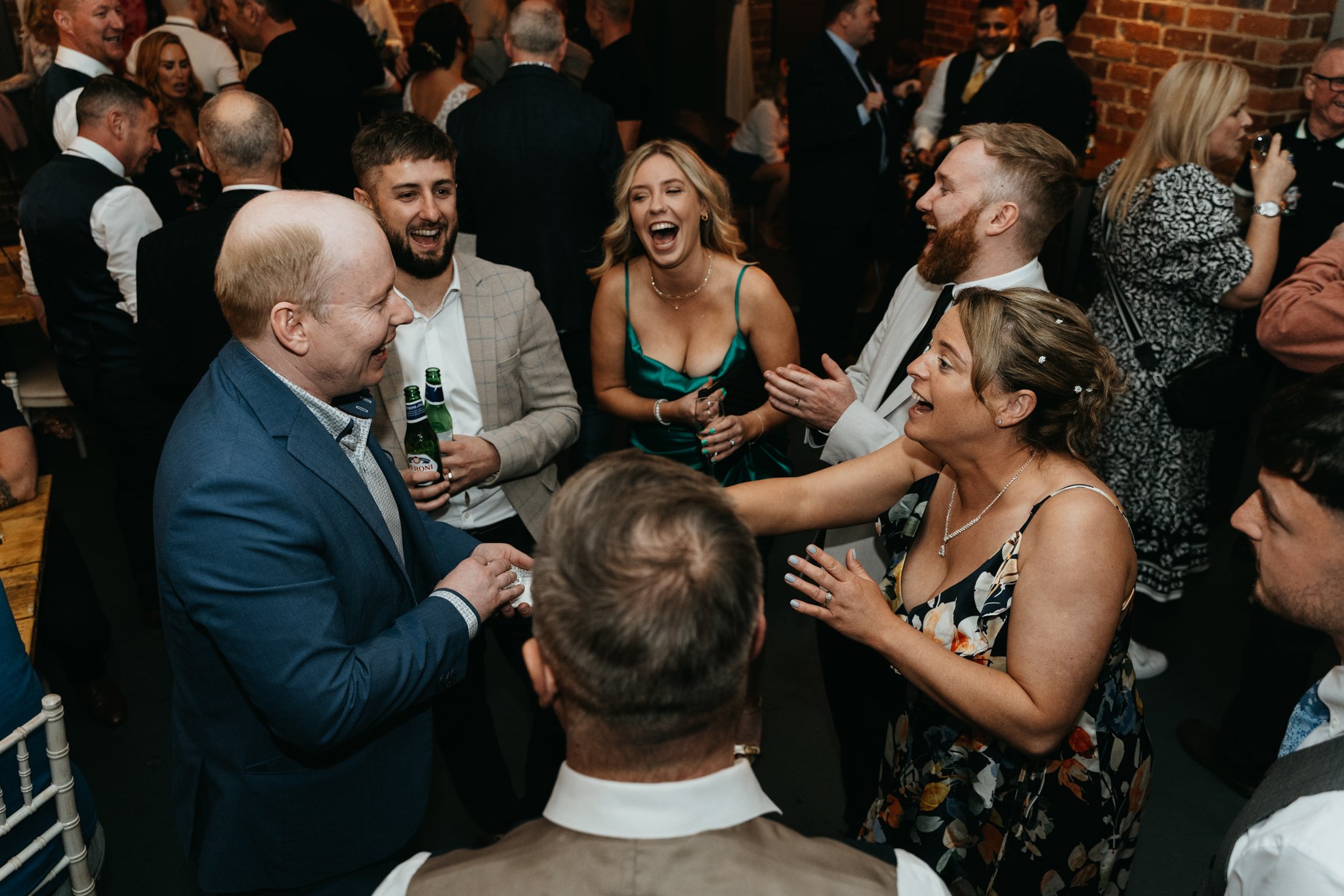 Group of people at a wedding reception celebrating and smiling, with some holding drinks, in a rustic indoor setting with exposed brick walls.