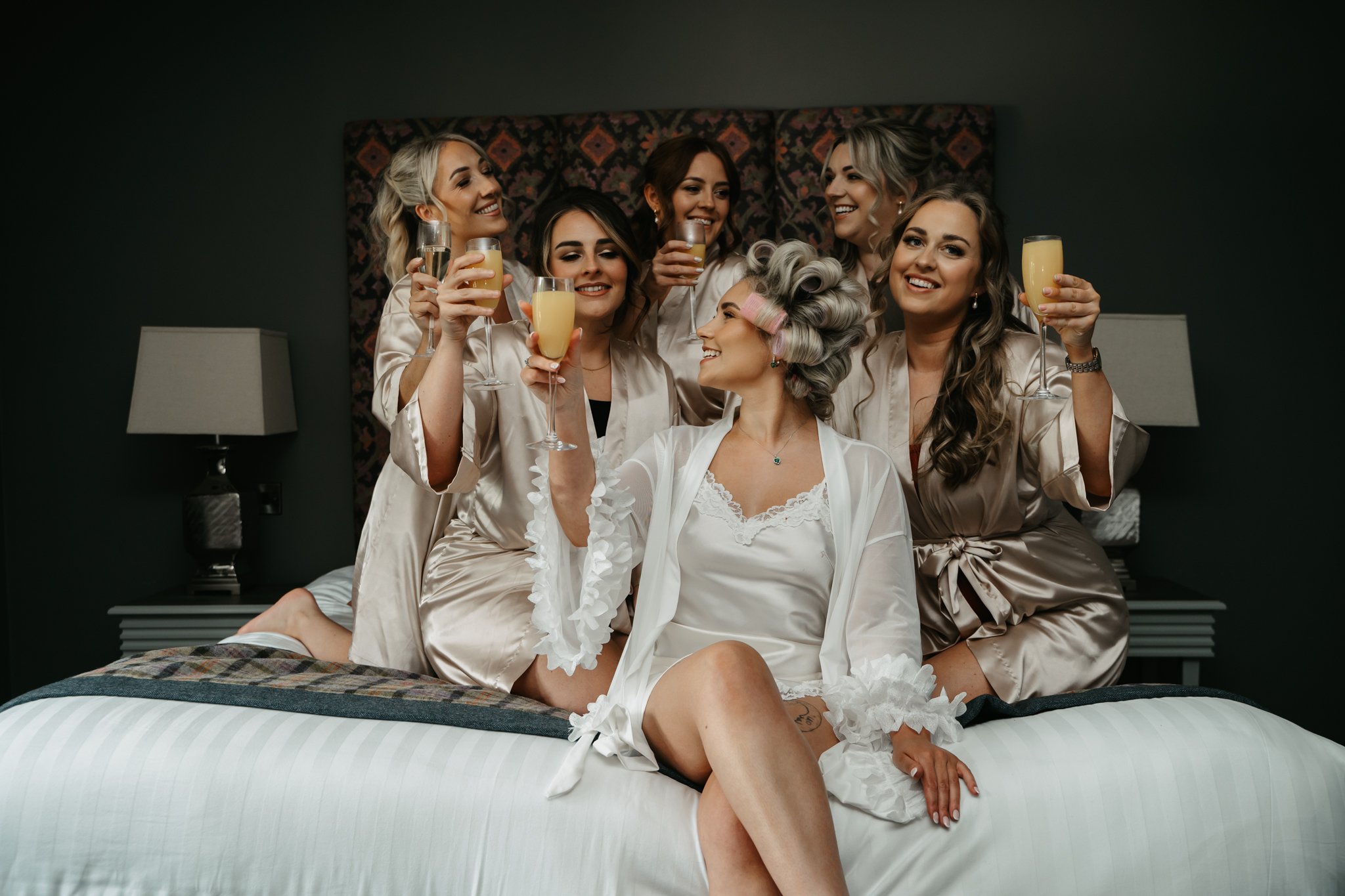 A bride and six bridesmaids in satin robes celebrating with champagne in a hotel room.