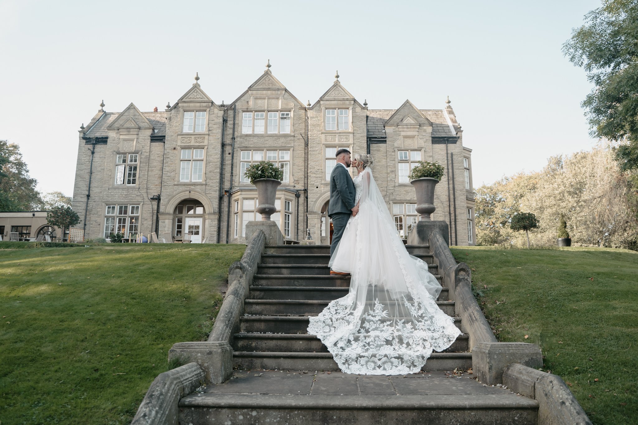 Bride and groom standing on outdoor stairs in front of a large historic mansion, holding hands and touching foreheads on a sunny day.
