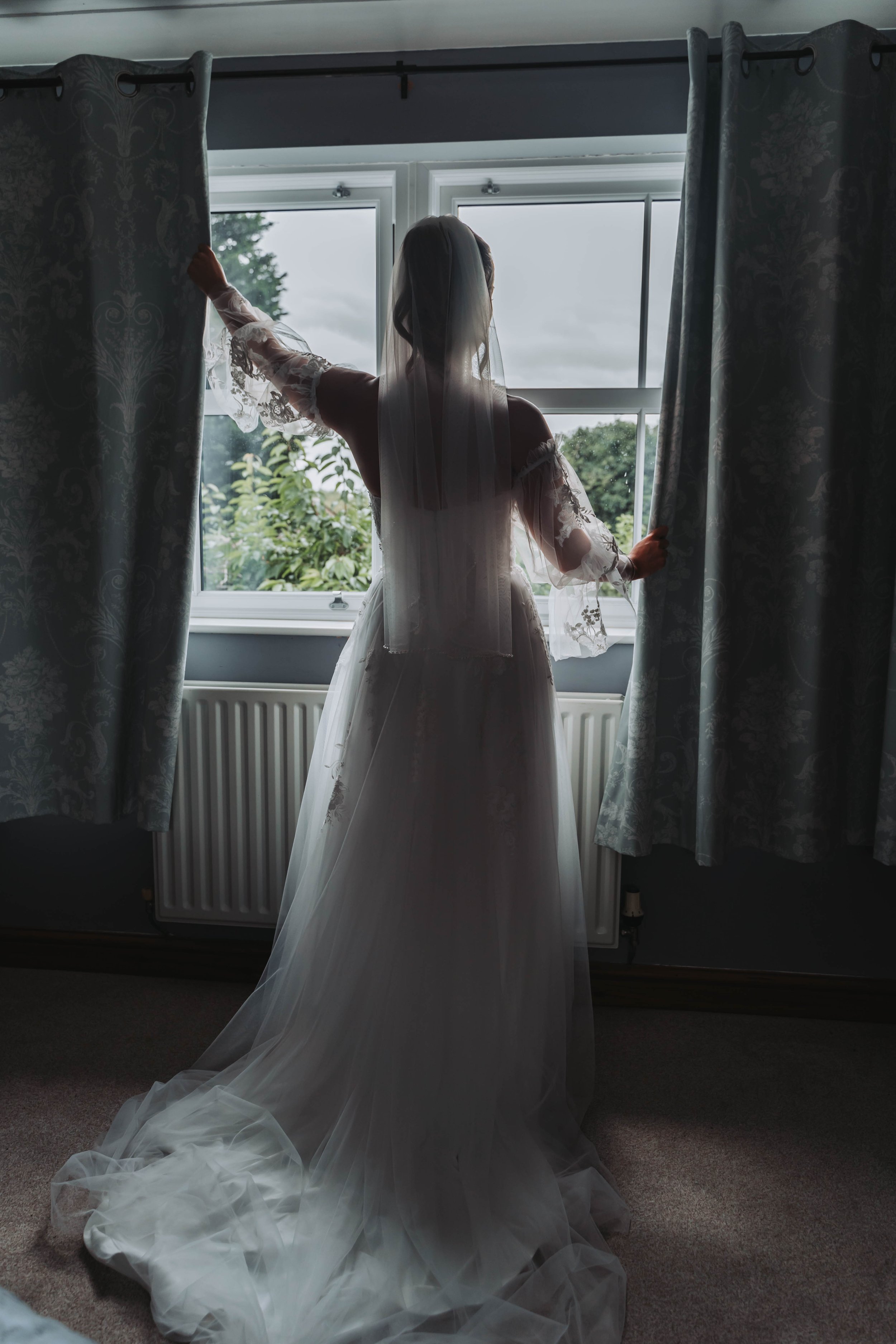 A bride in a wedding dress and veil standing by a window, holding back curtains to look outside.